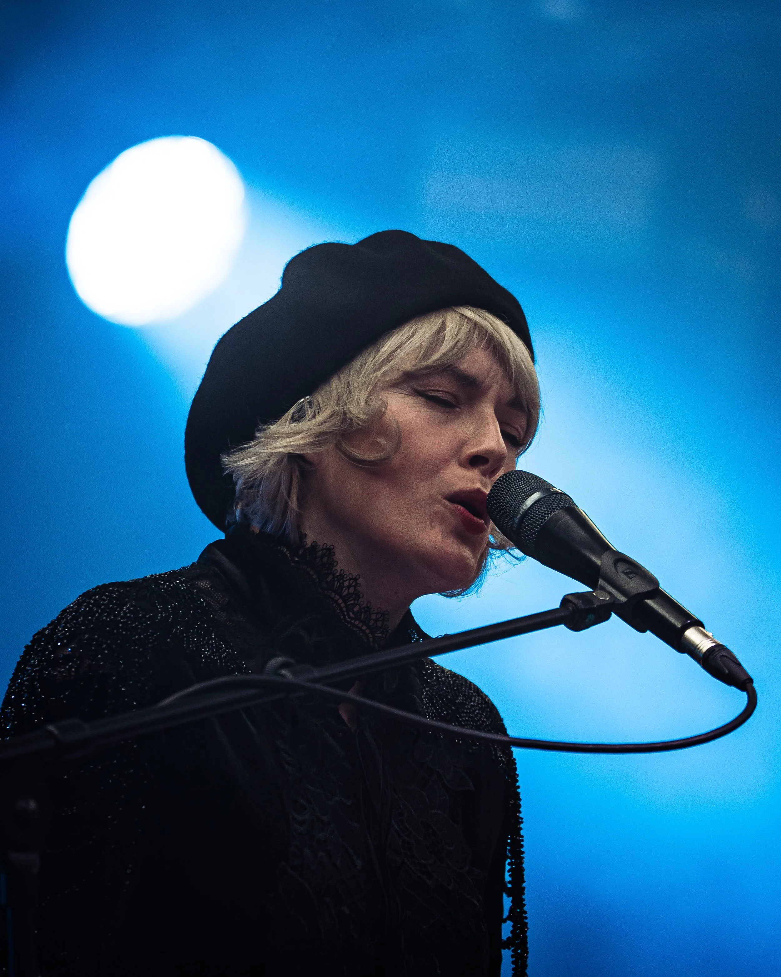 A person with blonde hair singing into a microphone, wearing a black hat and a black, sparkly top, against a blue background with a bright, out-of-focus light.