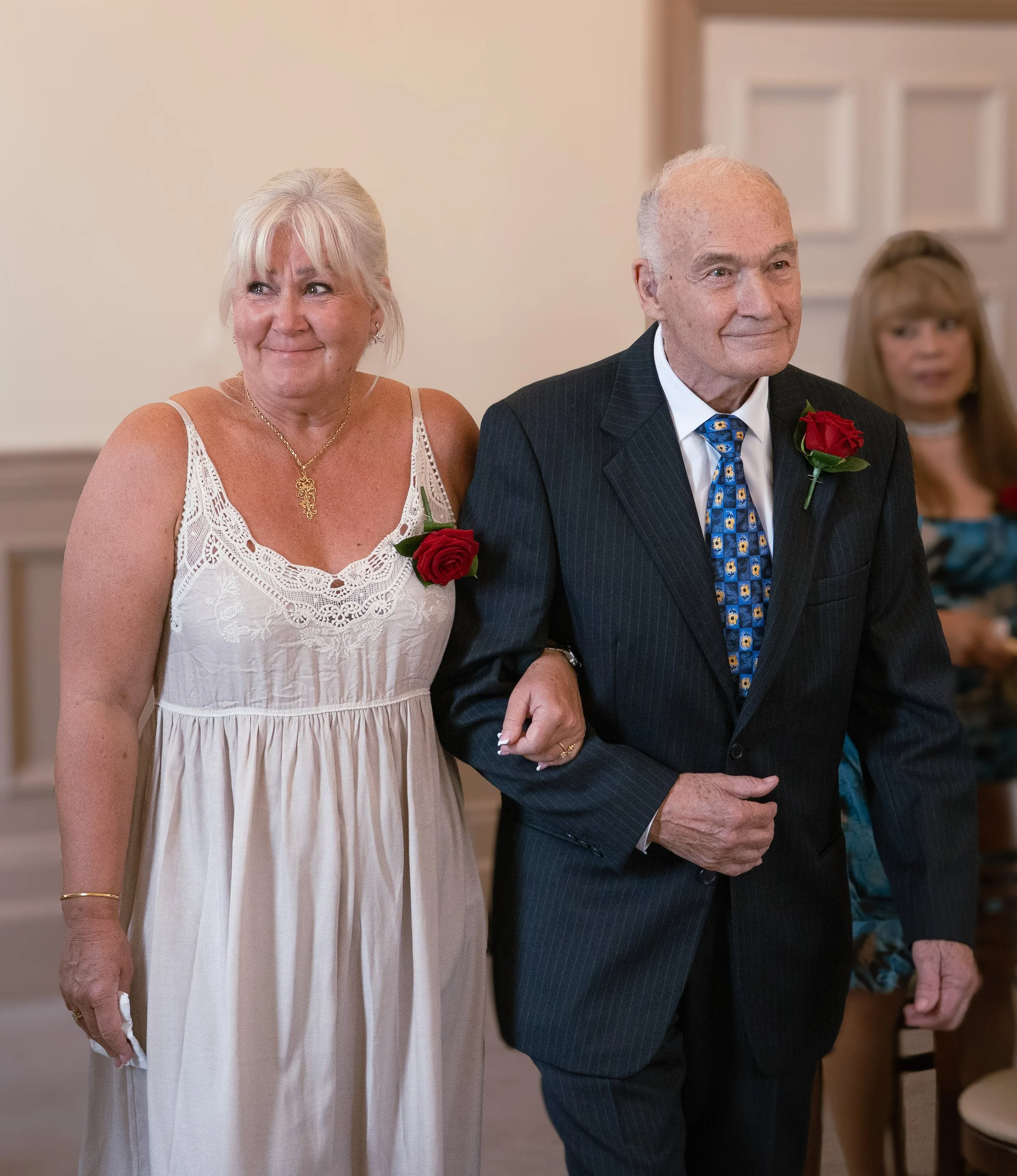 An elderly man dressed in a dark suit with a blue patterned tie walking arm-in-arm with a woman in a white sleeveless dress, both wearing red rose boutonnieres, at what appears to be a formal event or wedding.