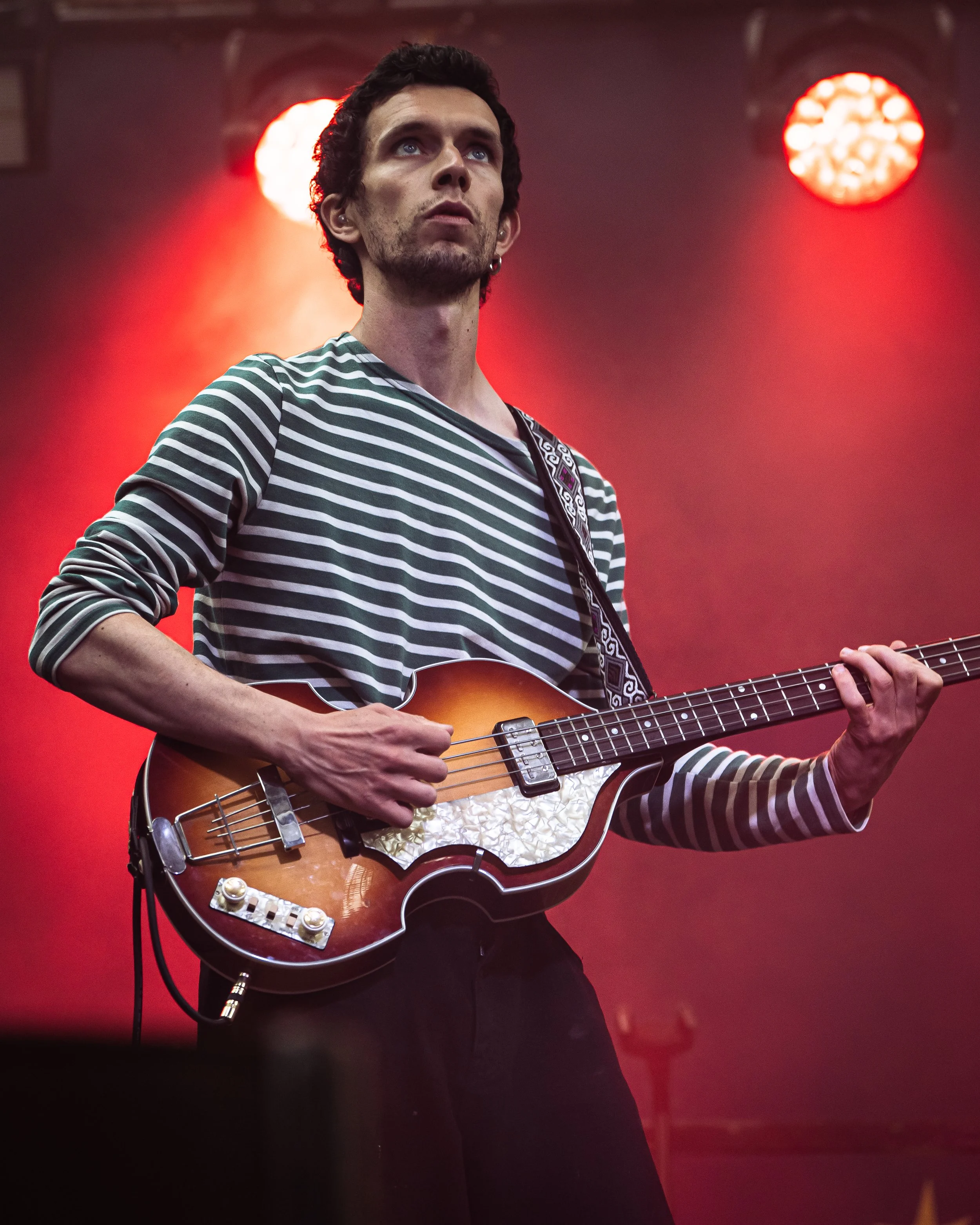 A man with dark curly hair and a beard playing an acoustic guitar on stage with red lighting in the background.