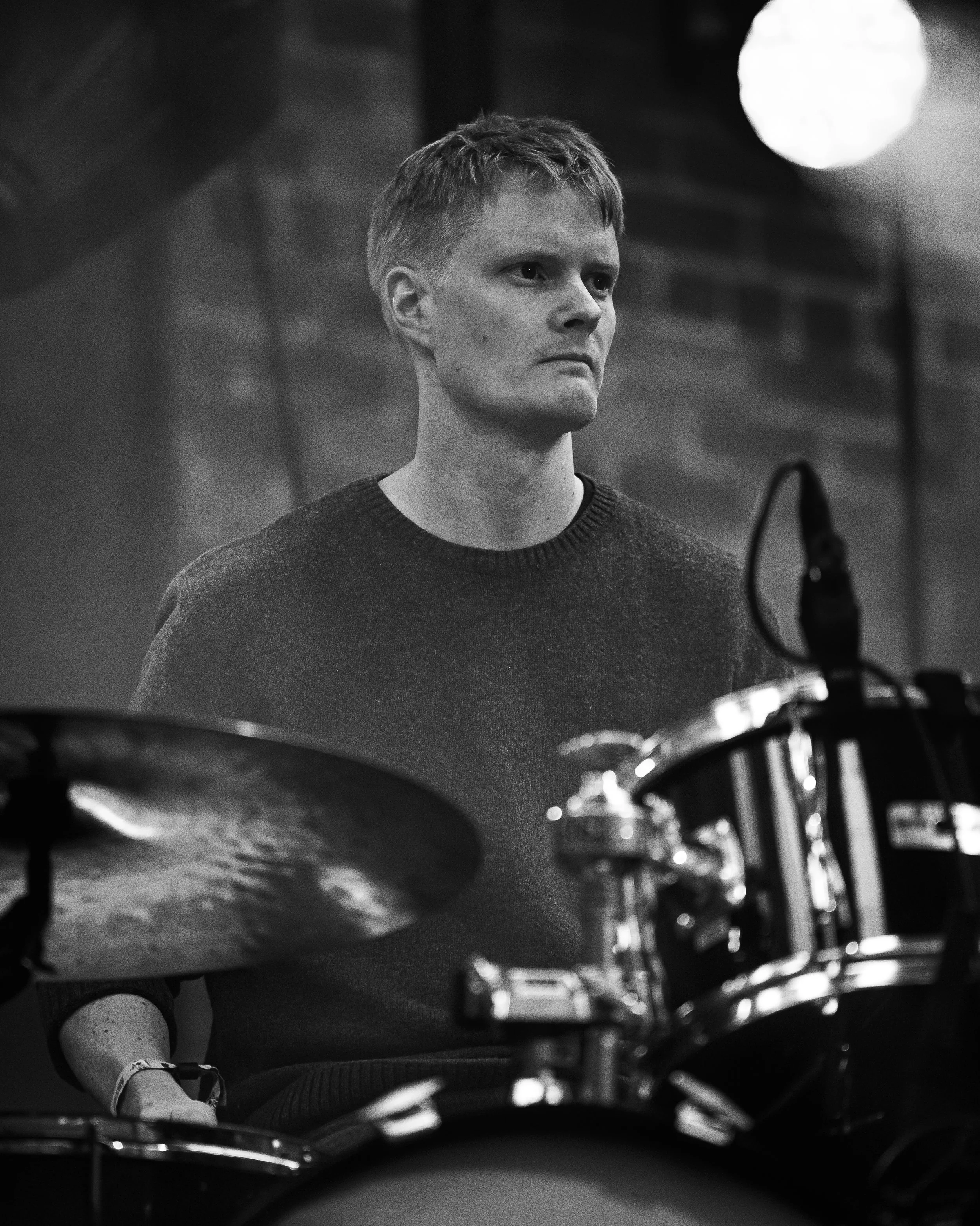 A person with short hair, wearing a dark sweater, standing behind drums and looking to the side, with a blurred background and microphone in the foreground.