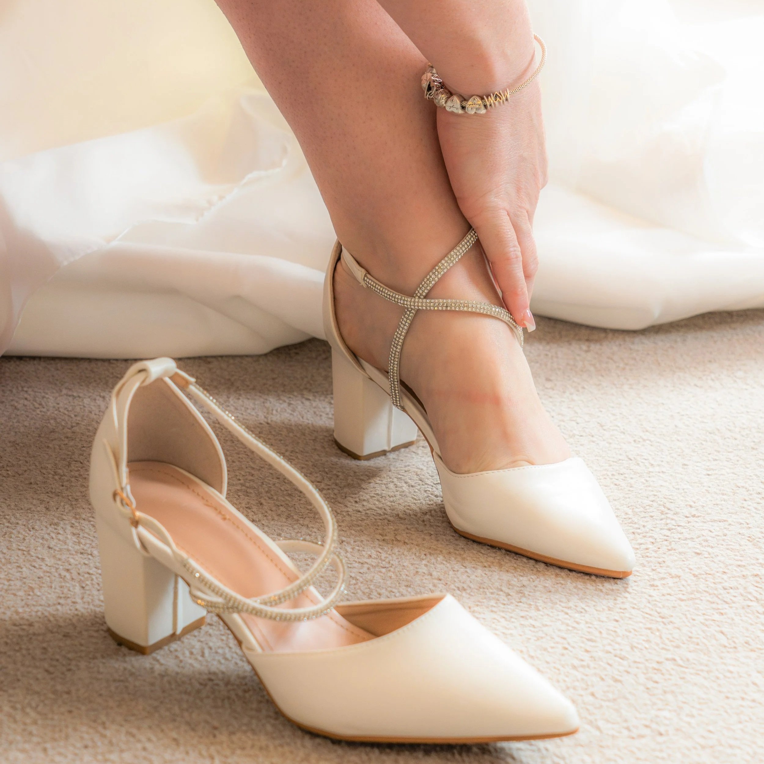 A woman in a white dress is adjusting and putting on a pair of cream-colored high-heeled shoes with ankle straps, while one shoe rests on the beige carpet.