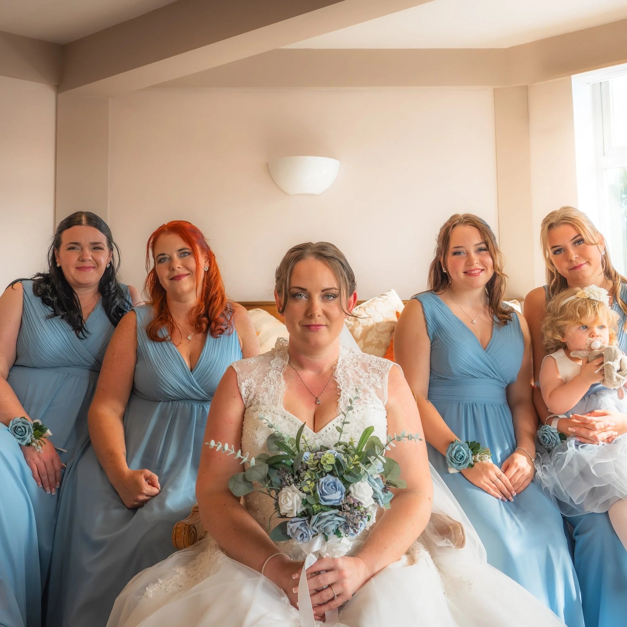 A bride in a wedding dress holding a bouquet, surrounded by her bridesmaids in blue dresses and a young girl in a blue dress with a pacifier, all sitting on a bed in a room with natural light.