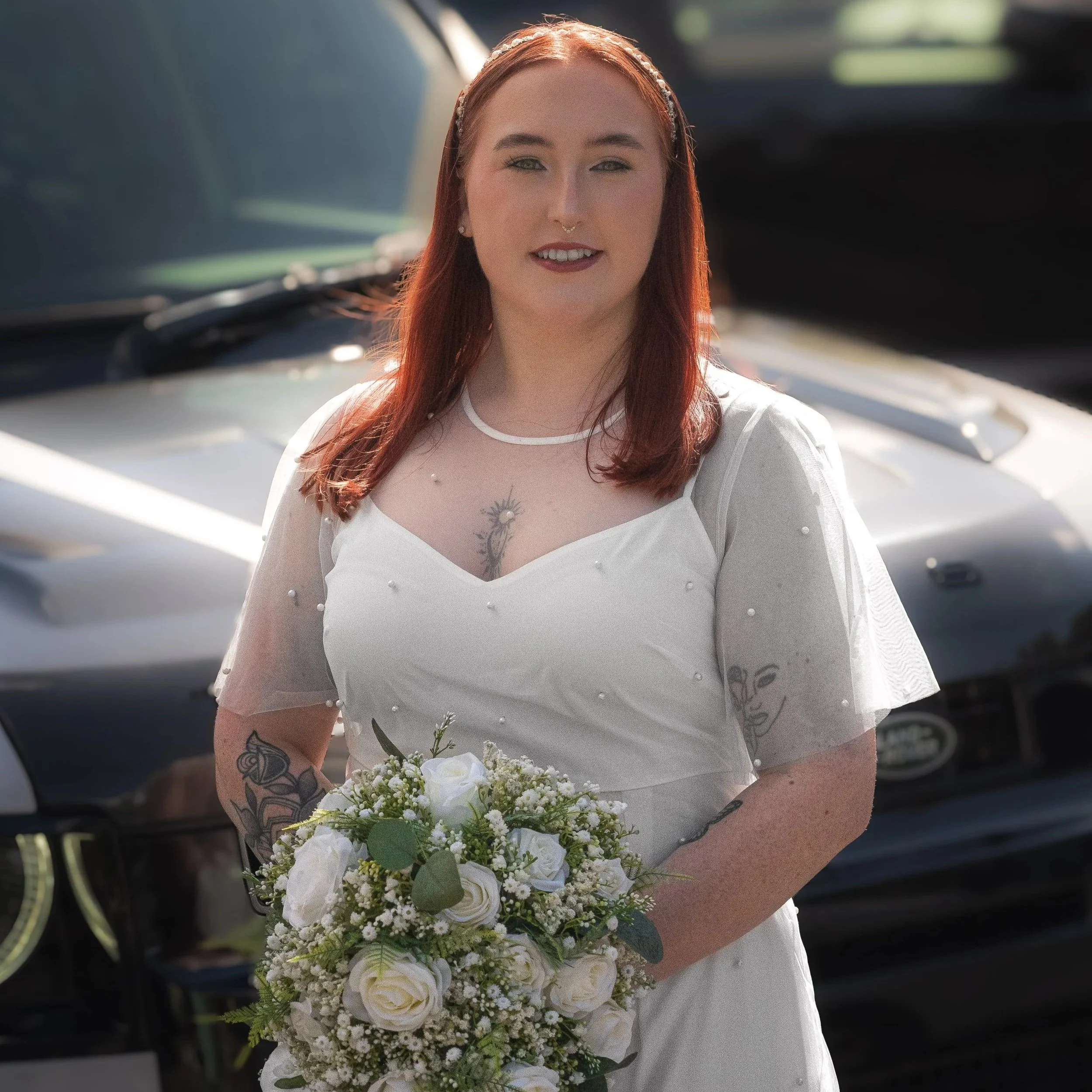 A young woman with red hair dressed in a white wedding gown holding a bouquet of white and green flowers standing outdoors in front of black cars.