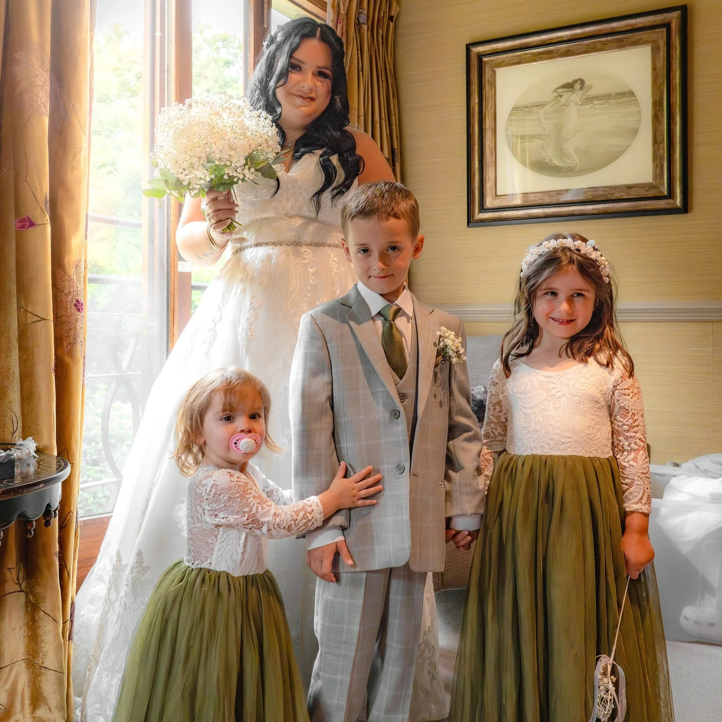 A bride in a white wedding dress holding a bouquet of flowers stands behind a group of four children, three girls and one boy, dressed formally. The children are in a room with beige walls and a framed picture of a woman on the wall, near a window wi