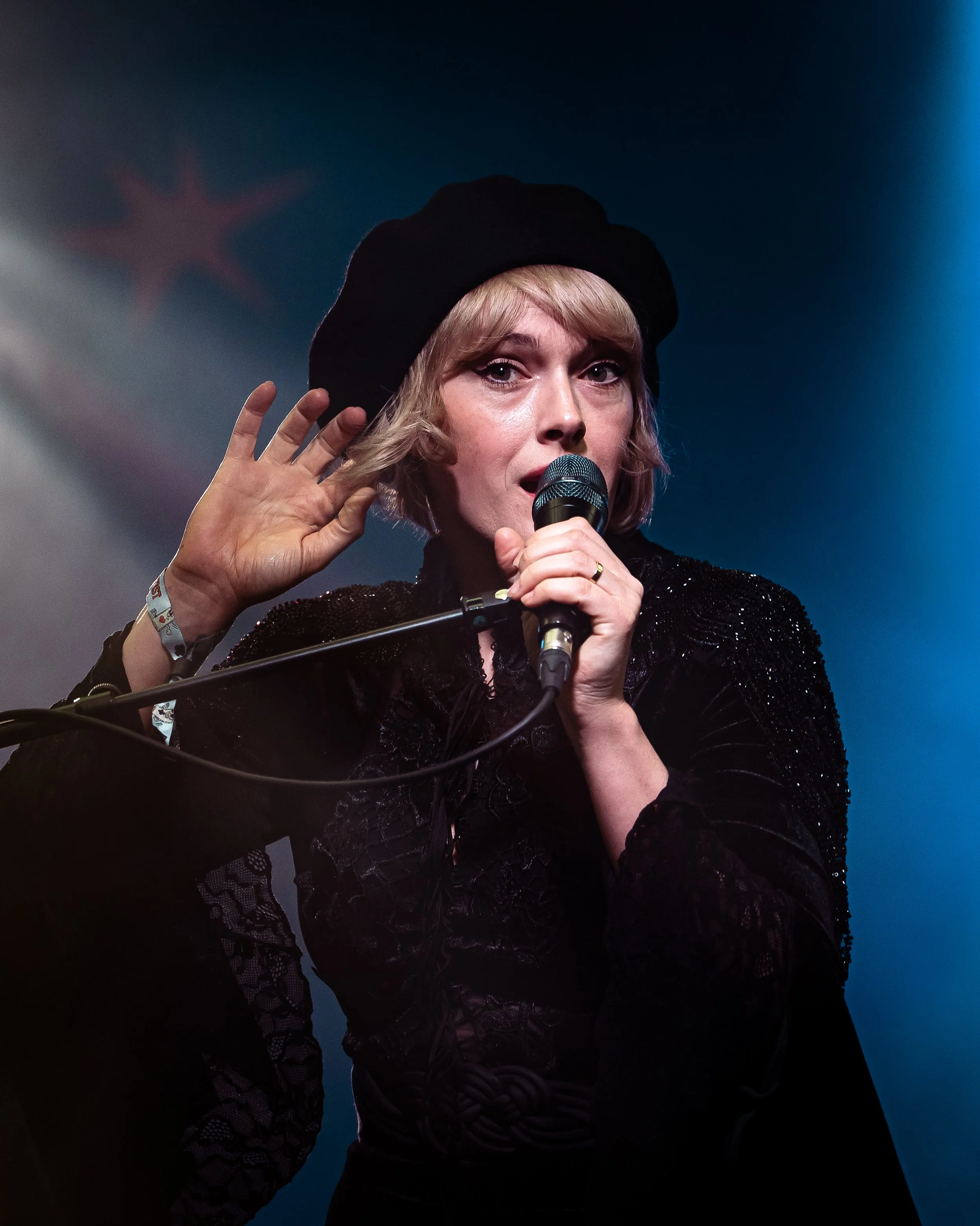 A woman with short blonde hair and light makeup holds a microphone and appears to be speaking or singing on stage. She is wearing a black hat and a black sequined outfit, and is raising her left hand near her ear. The background is dark with a subtle
