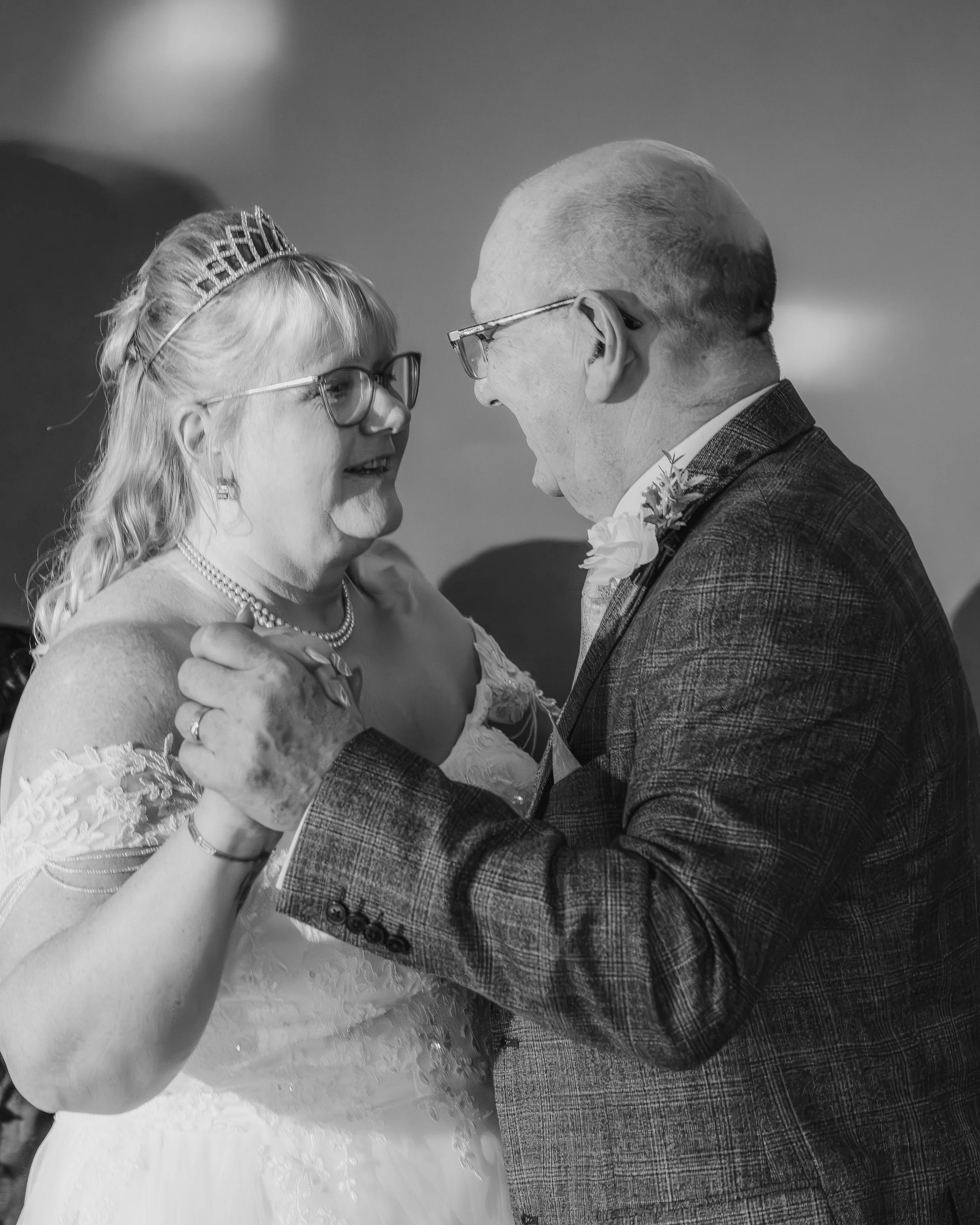 A bride and groom dancing at their wedding, smiling and looking at each other, with the bride wearing glasses and a tiara, and the groom wearing glasses and a boutonniere, in a black-and-white photo.