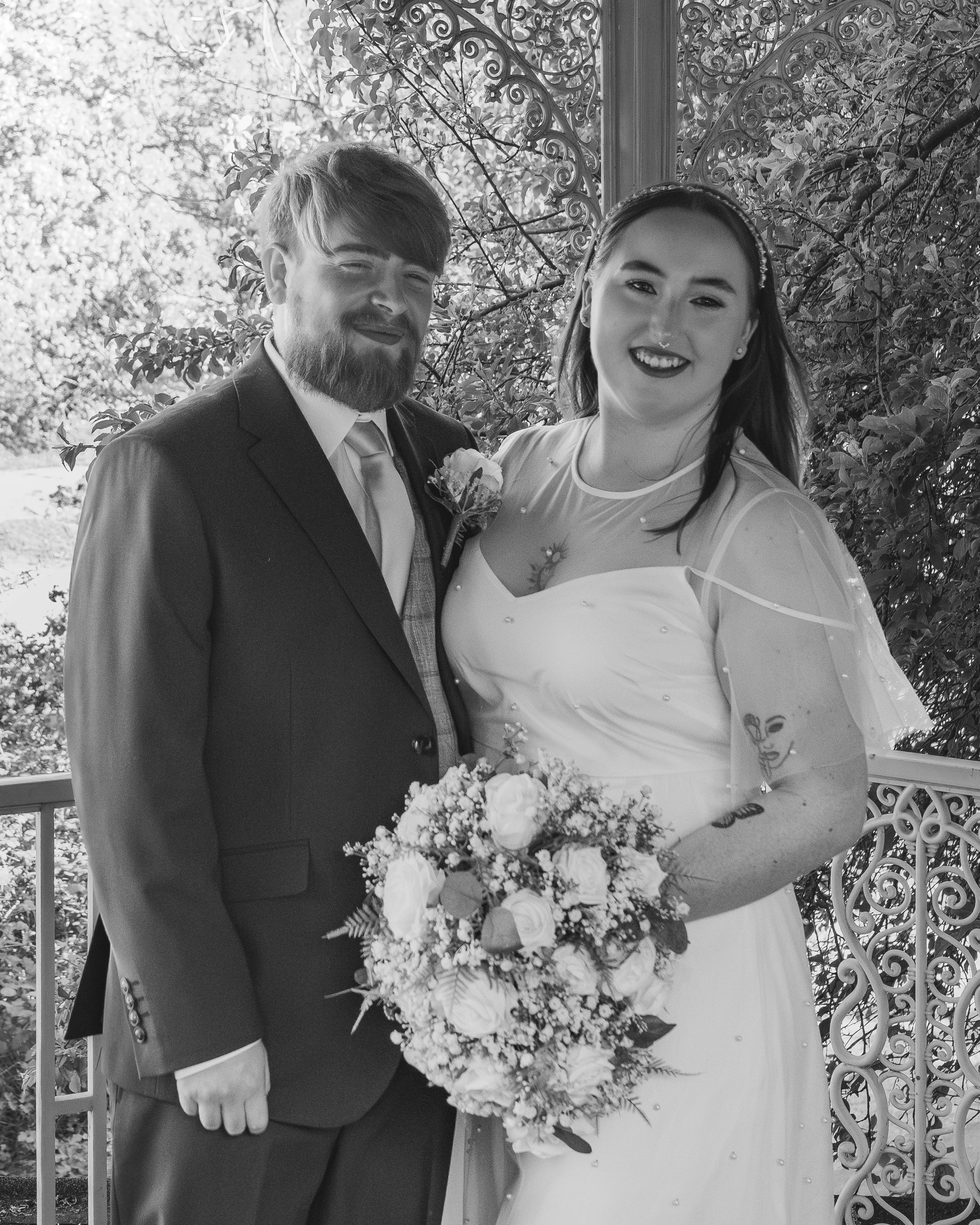 A bride and groom on their wedding day standing outdoors. The bride is holding a bouquet of flowers and wearing a wedding dress. The groom is dressed in a suit and tie. They are smiling and standing close together.