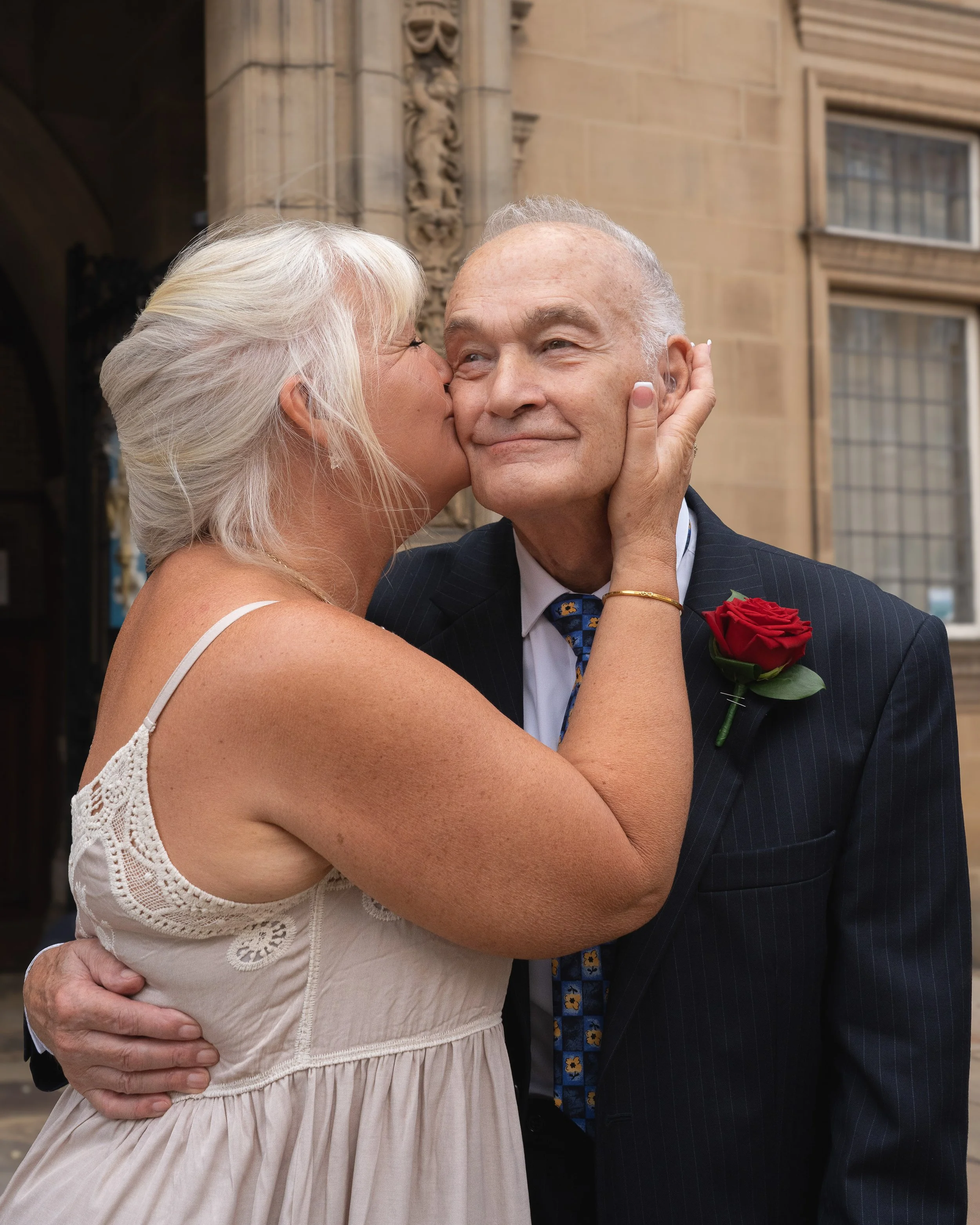 A woman with blonde hair in a white dress is kissing a man with gray hair in a navy suit on the cheek outside a building with stone walls and large windows.