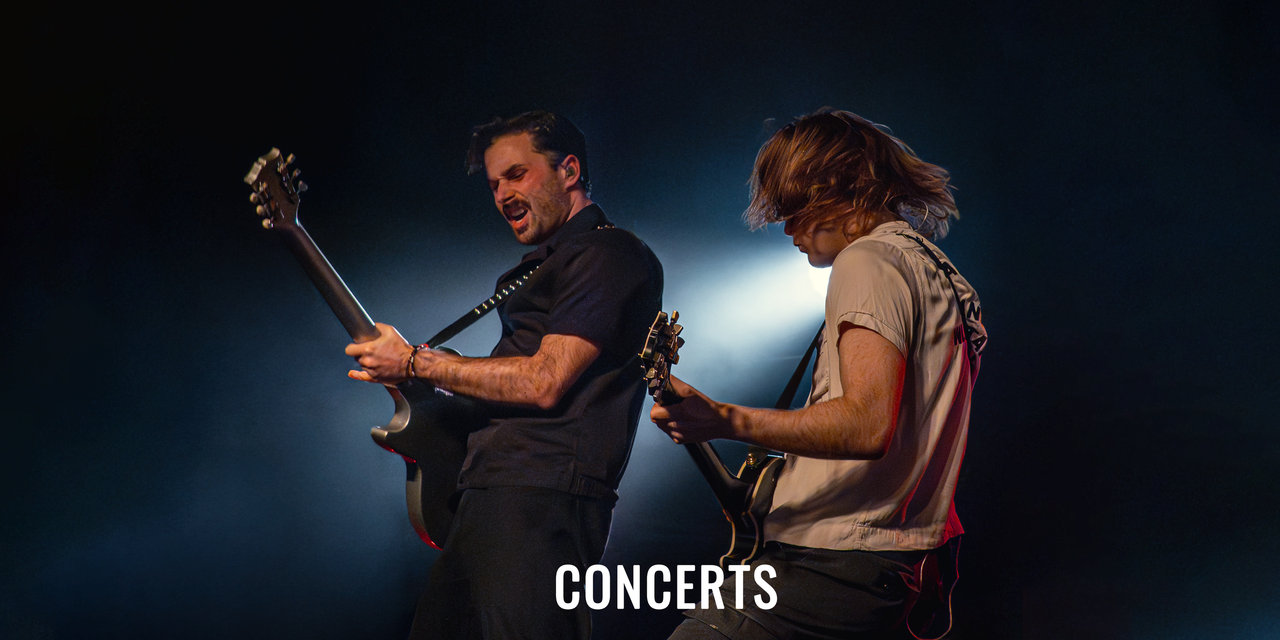 Two male musicians playing electric guitars on stage during a concert with a bright light behind them, creating a dramatic scene.