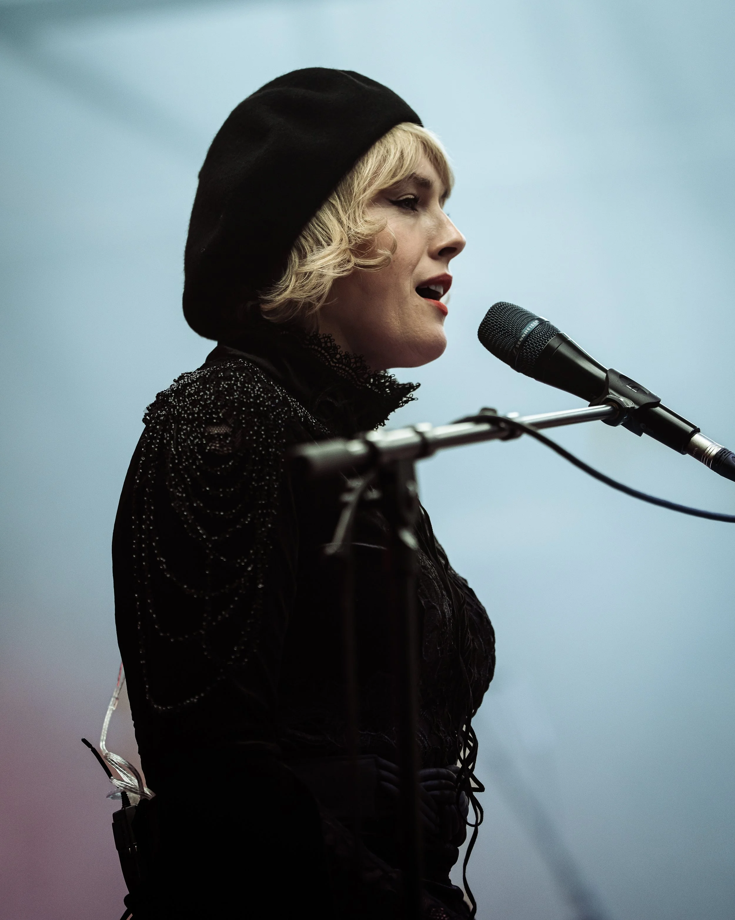 A woman singing into a microphone, wearing a black hat and a black outfit, with a blurred light blue background.