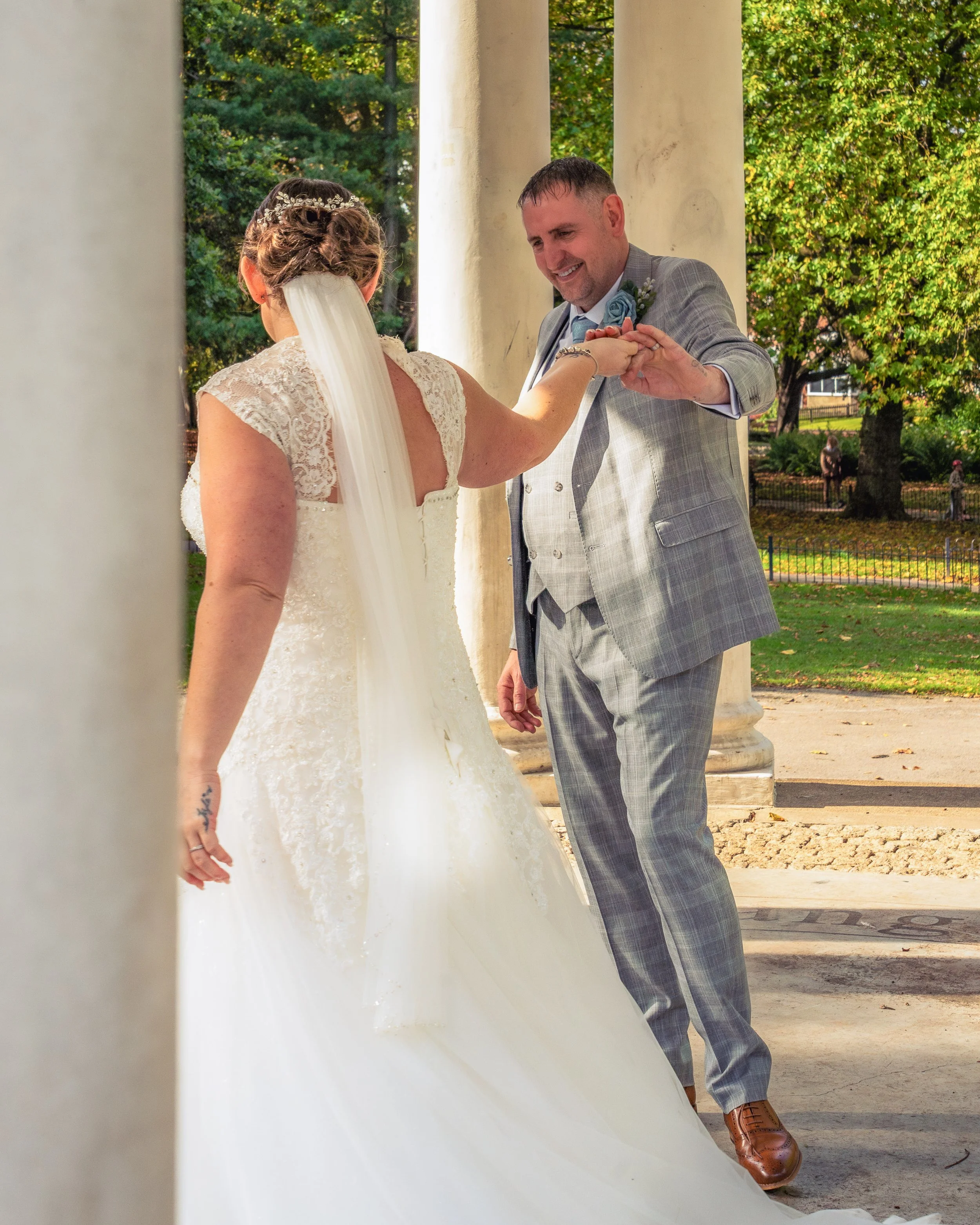 A bride and groom exchanging wedding rings outdoors under classical columns with green trees in the background.