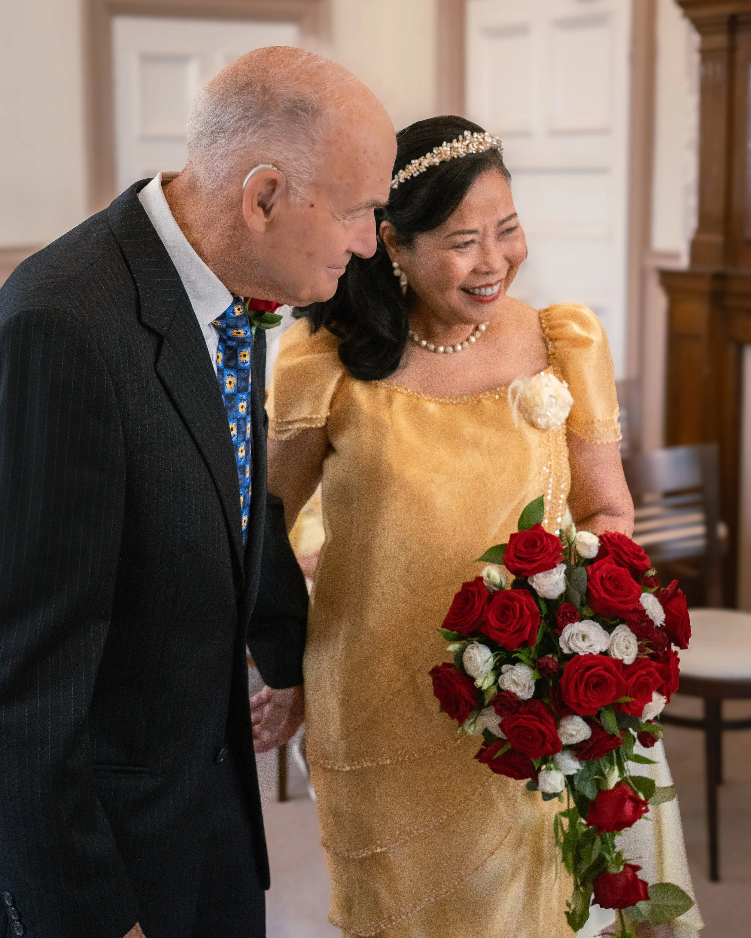 An elderly man and woman smiling and holding hands, with the woman holding a bouquet of red and white roses, in a room with wooden furnishings.