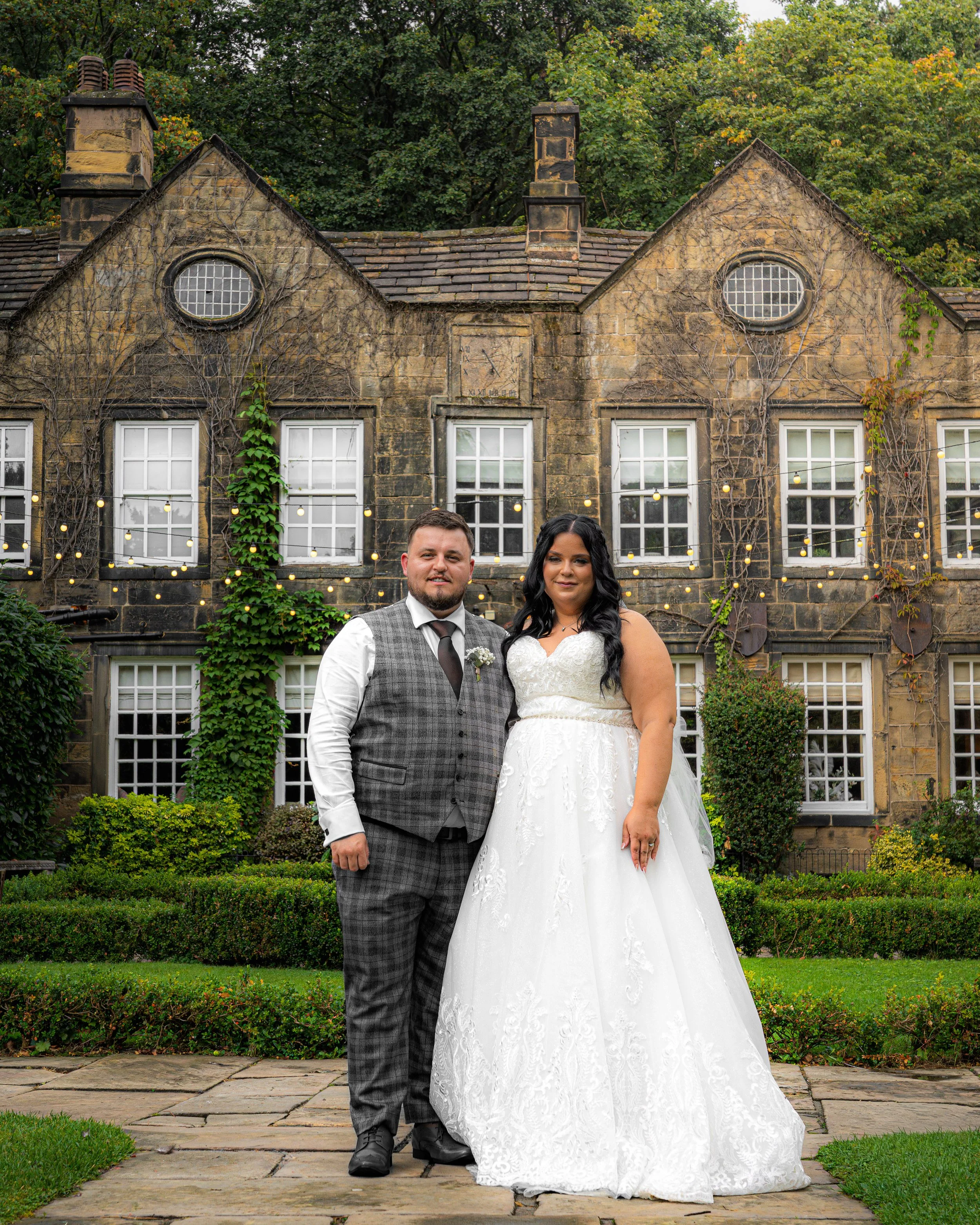A newlywed couple standing outside in front of an old stone building decorated with string lights and ivy, with lush greenery and a manicured garden surrounding them.