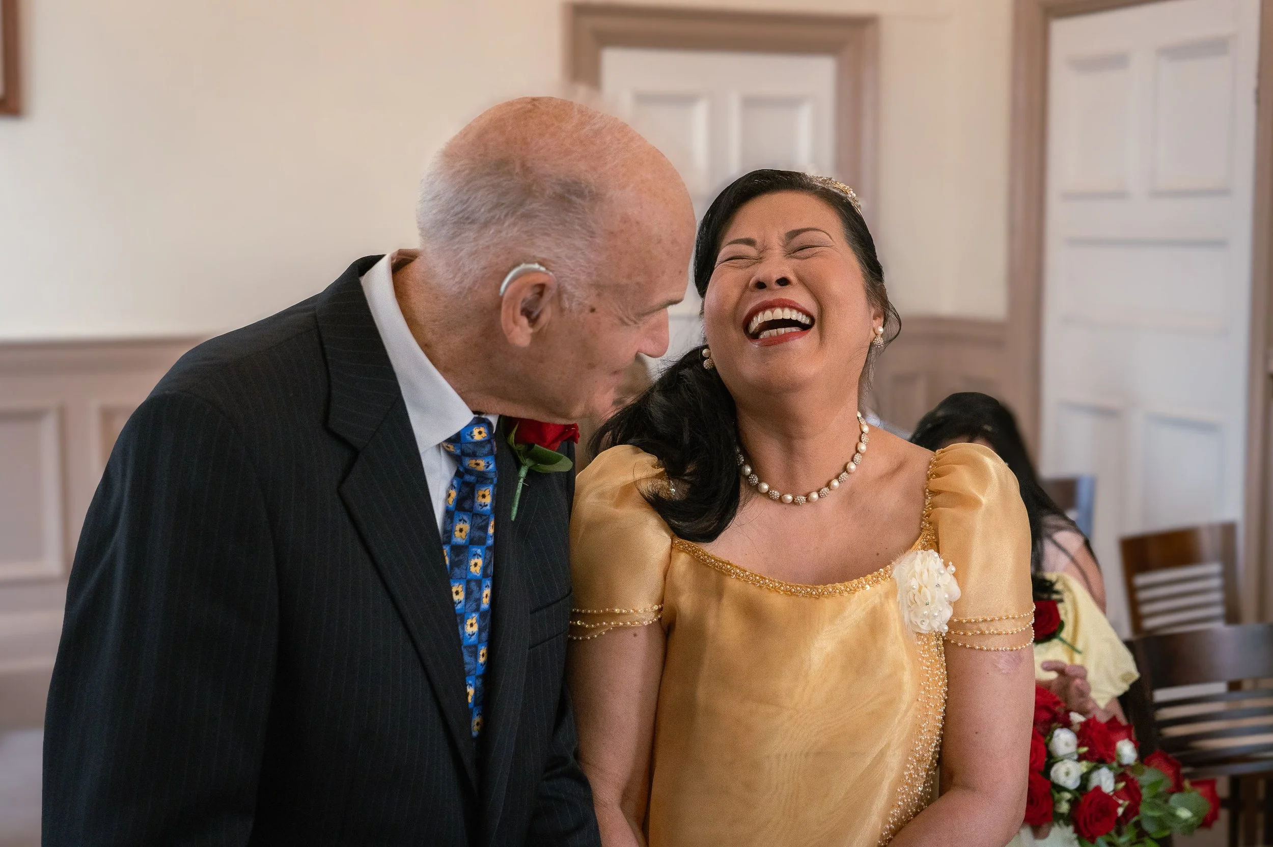 An elderly man and woman sharing a joyful moment at a wedding, with the woman smiling widely and the man looking at her affectionately.