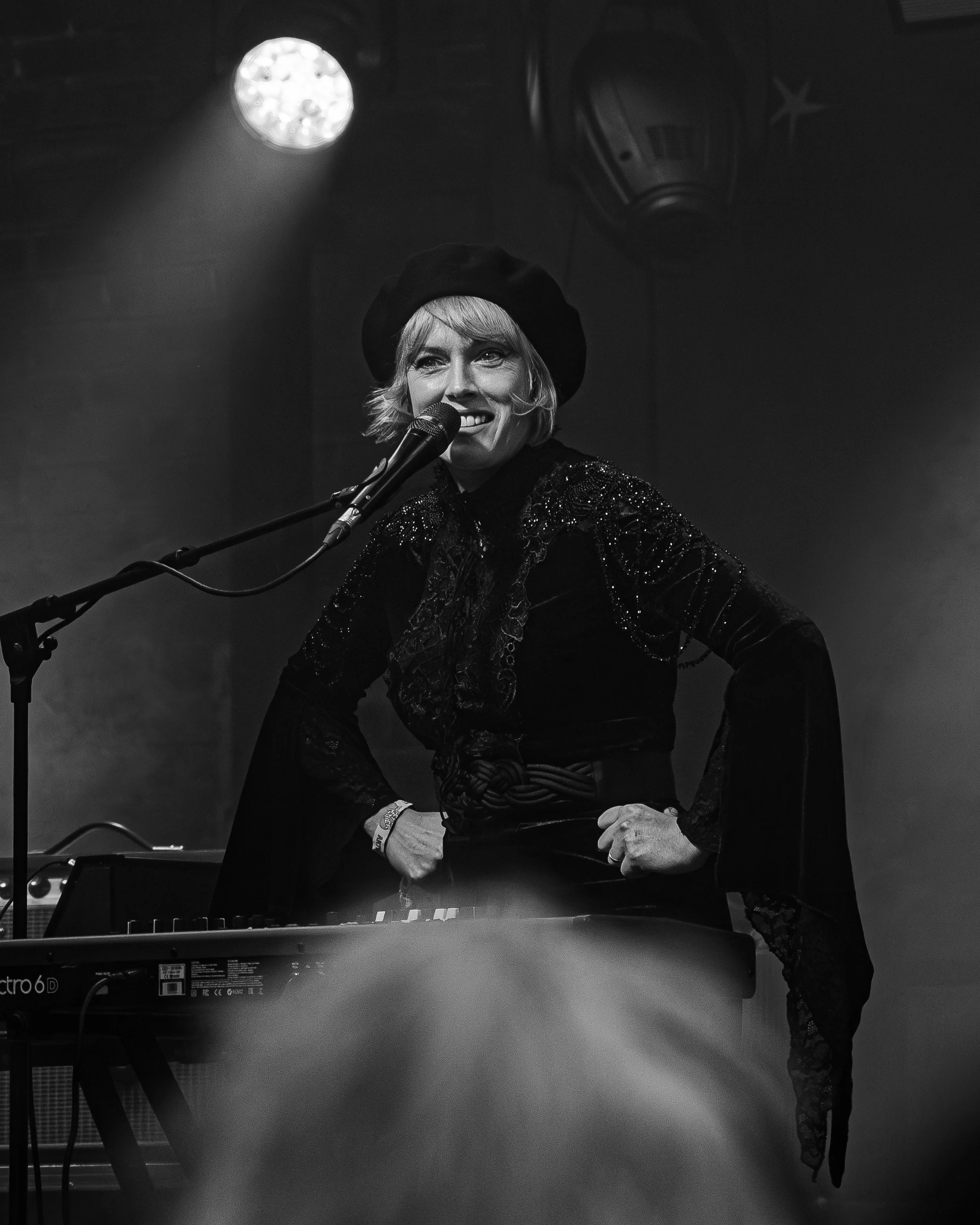 A woman smiling while singing into a microphone on stage, wearing a black beret and a shiny black jacket, with stage lights overhead.
