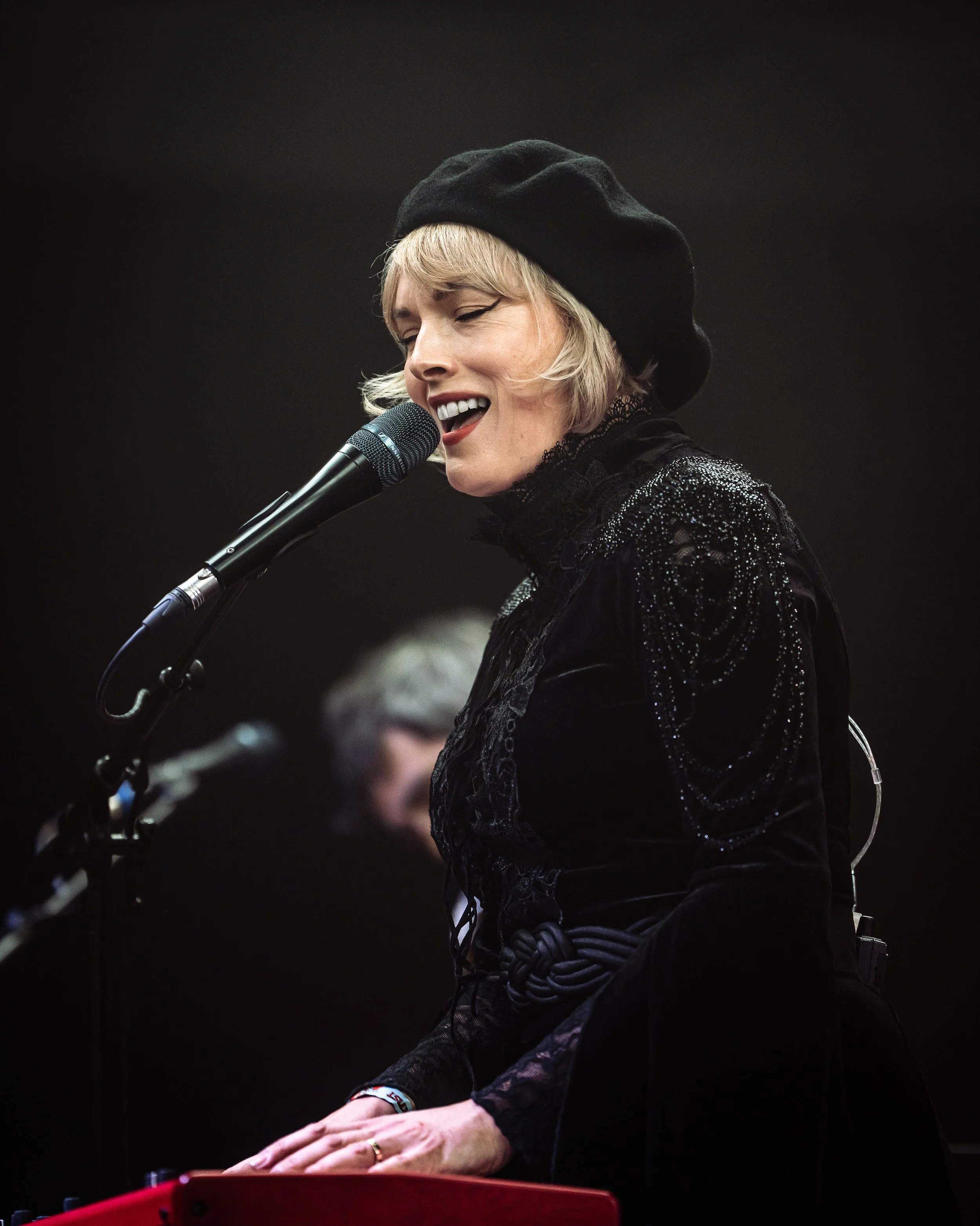 A woman with short blonde hair, wearing a black beret and black lace dress, is singing into a microphone, with her eyes closed and a smile on her face.