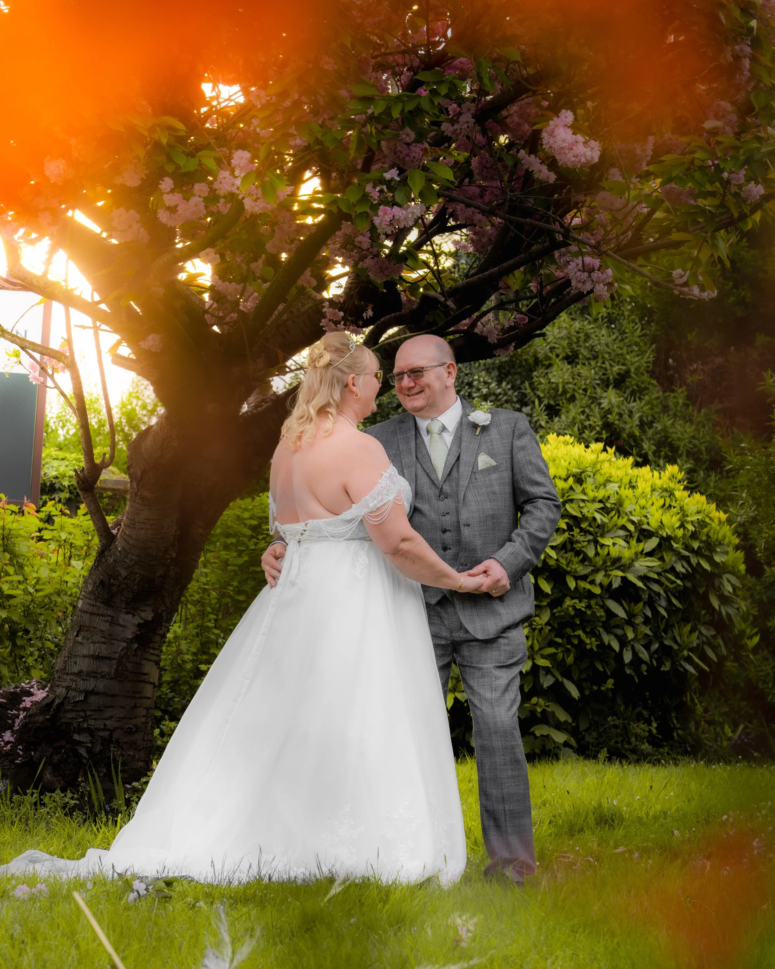 A bride and groom holding hands and smiling at each other outside, under a blooming pink cherry blossom tree during their wedding.