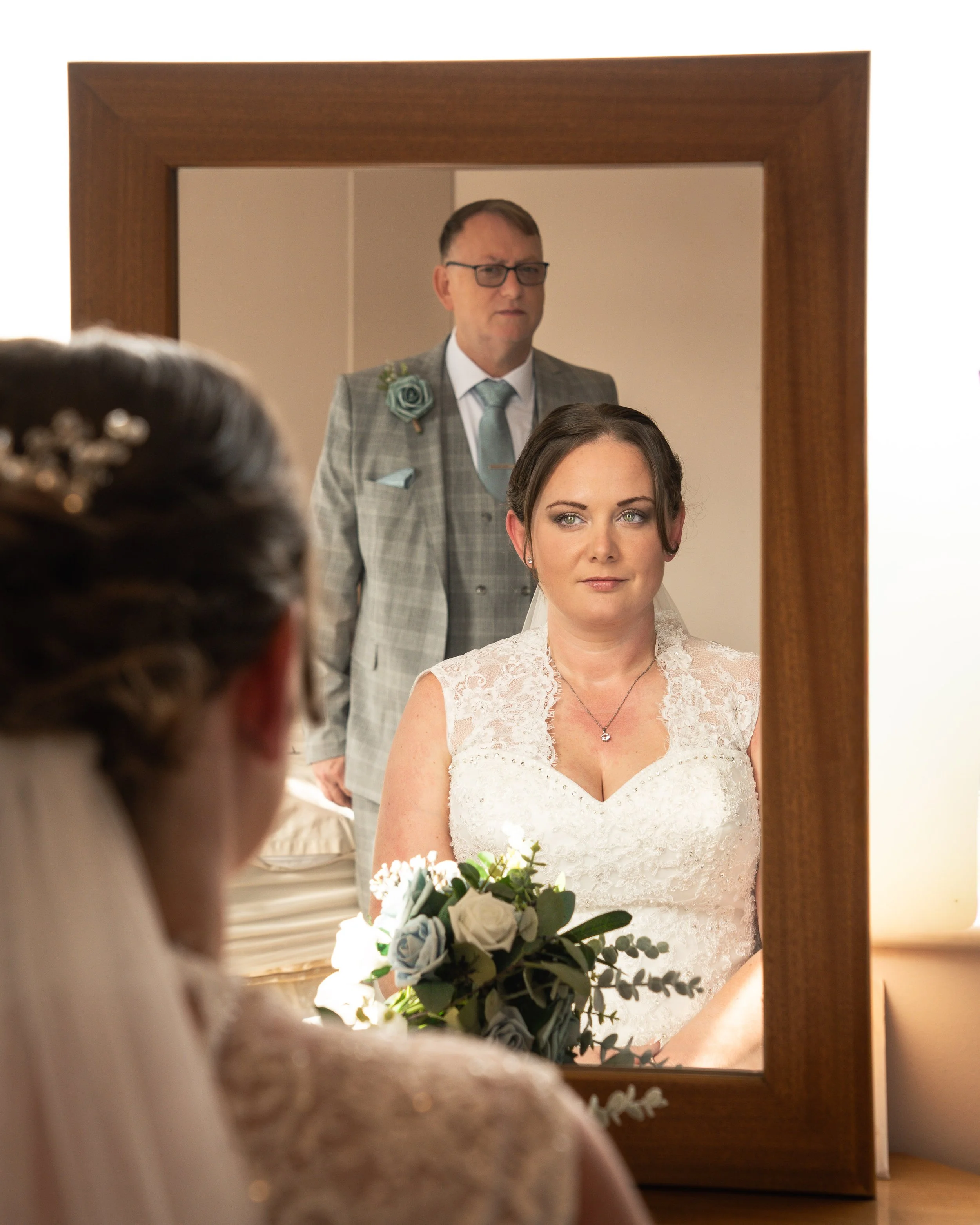 Bride in a white lace dress looking into a mirror with the groom in a plaid suit reflected behind her at a wedding preparation.