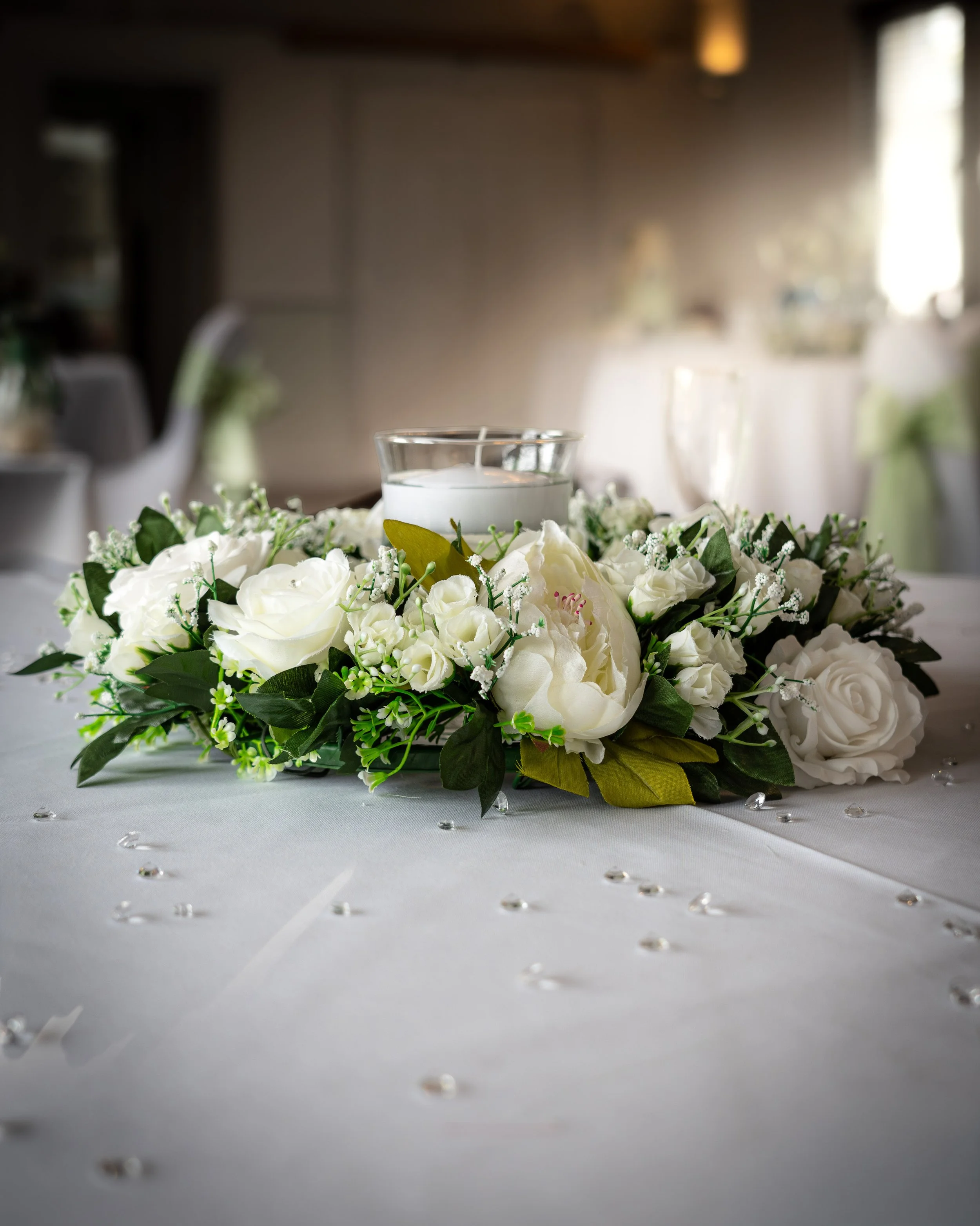 A floral centerpiece with white roses and greenery on a table, decorated with scattered clear rhinestones, and a glass candle holder with a white candle behind the flowers in a decorated setting.