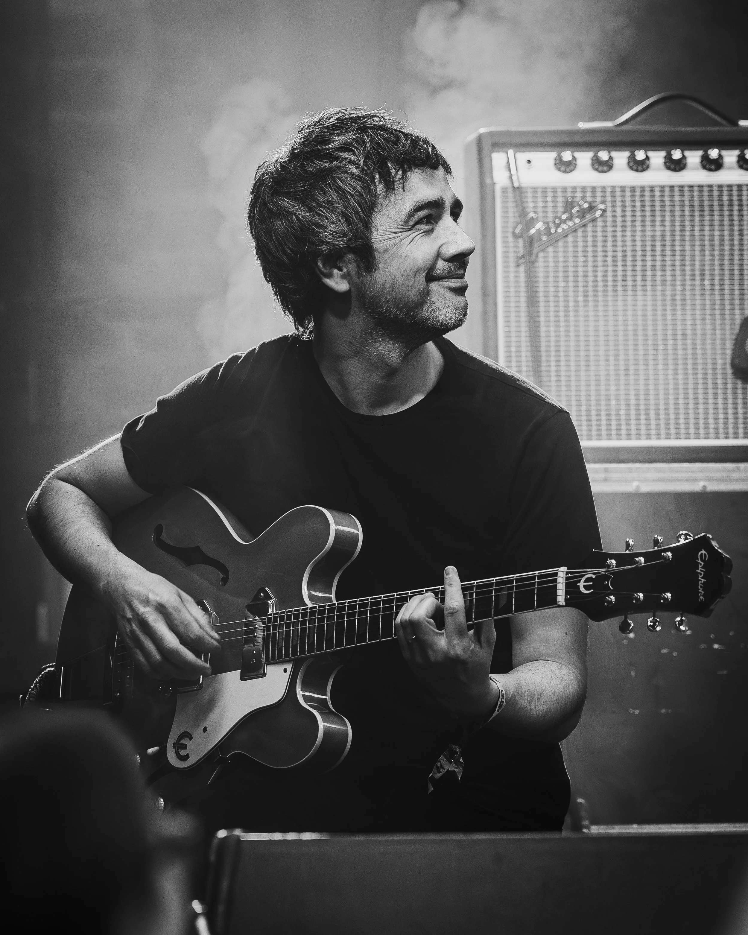 Black and white photo of a man smiling and playing an electric guitar in a music studio, with a Fender amplifier in the background.