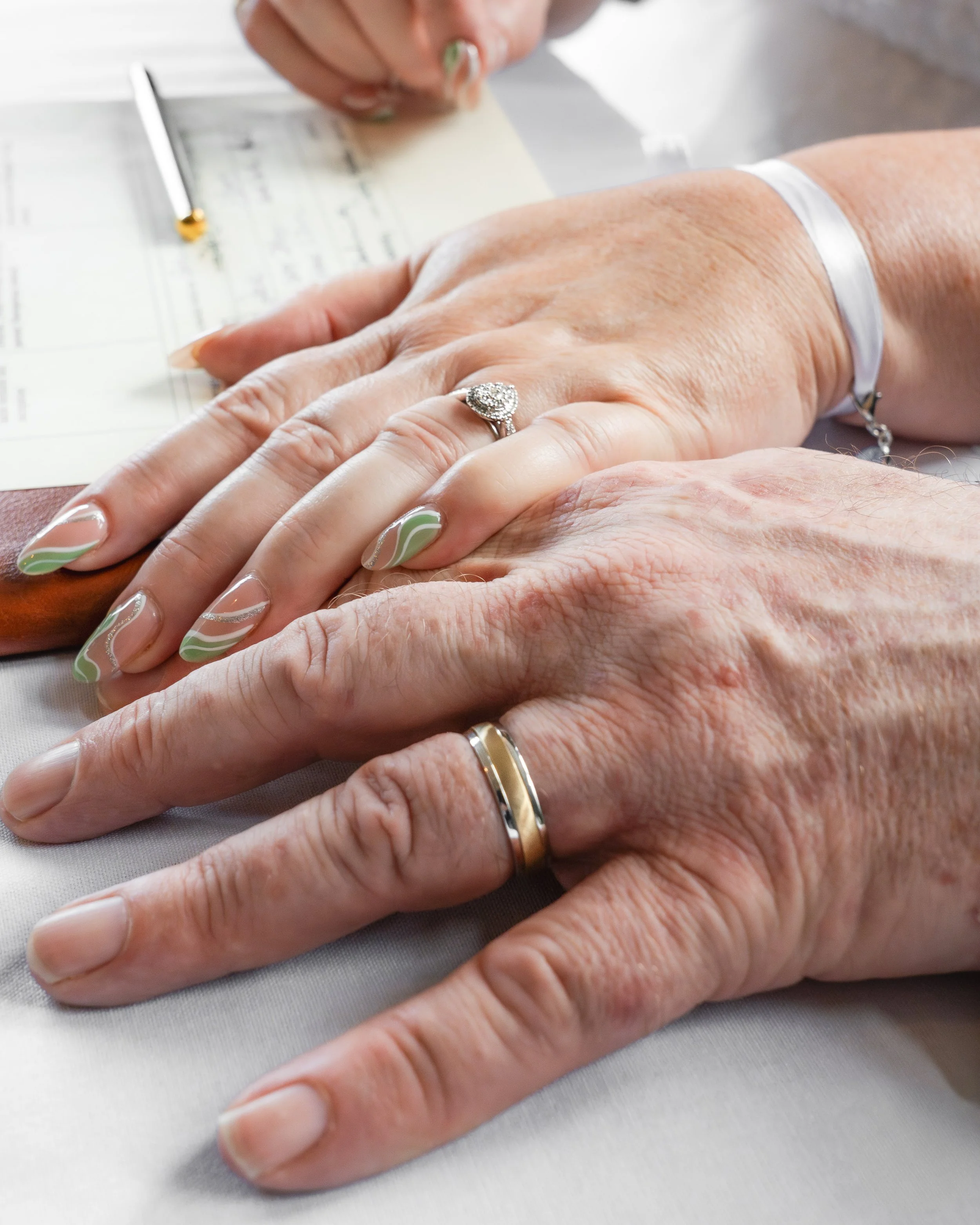 Close-up of two hands, one of an older person and one of a younger person, showing wedding rings, with pink and green nail polish on the younger person's nails, on a table with documents in the background.