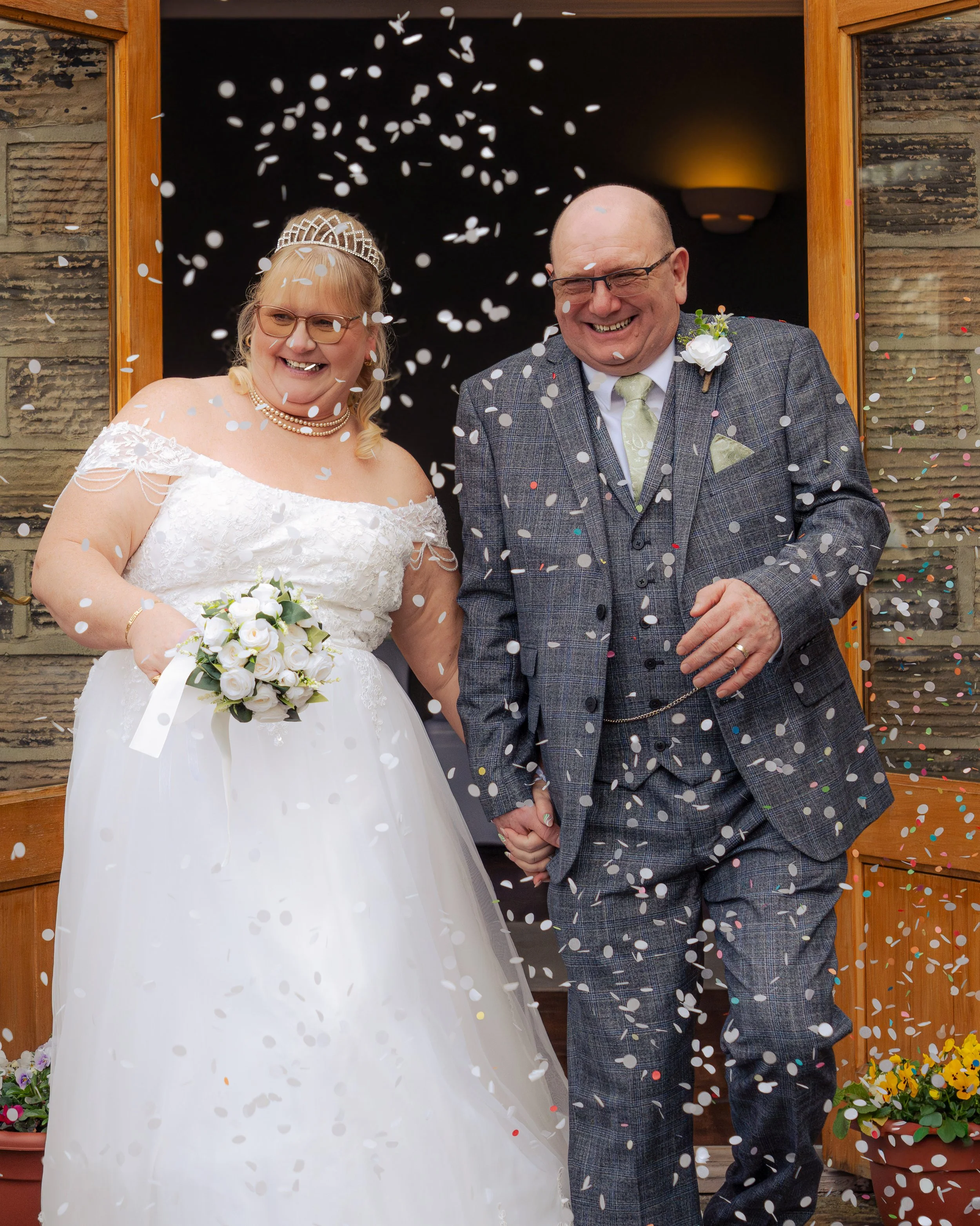 A newlywed couple, the bride in a white wedding dress holding a bouquet and the groom in a gray plaid suit, standing outside their wedding venue as confetti falls around them, both smiling and holding hands.