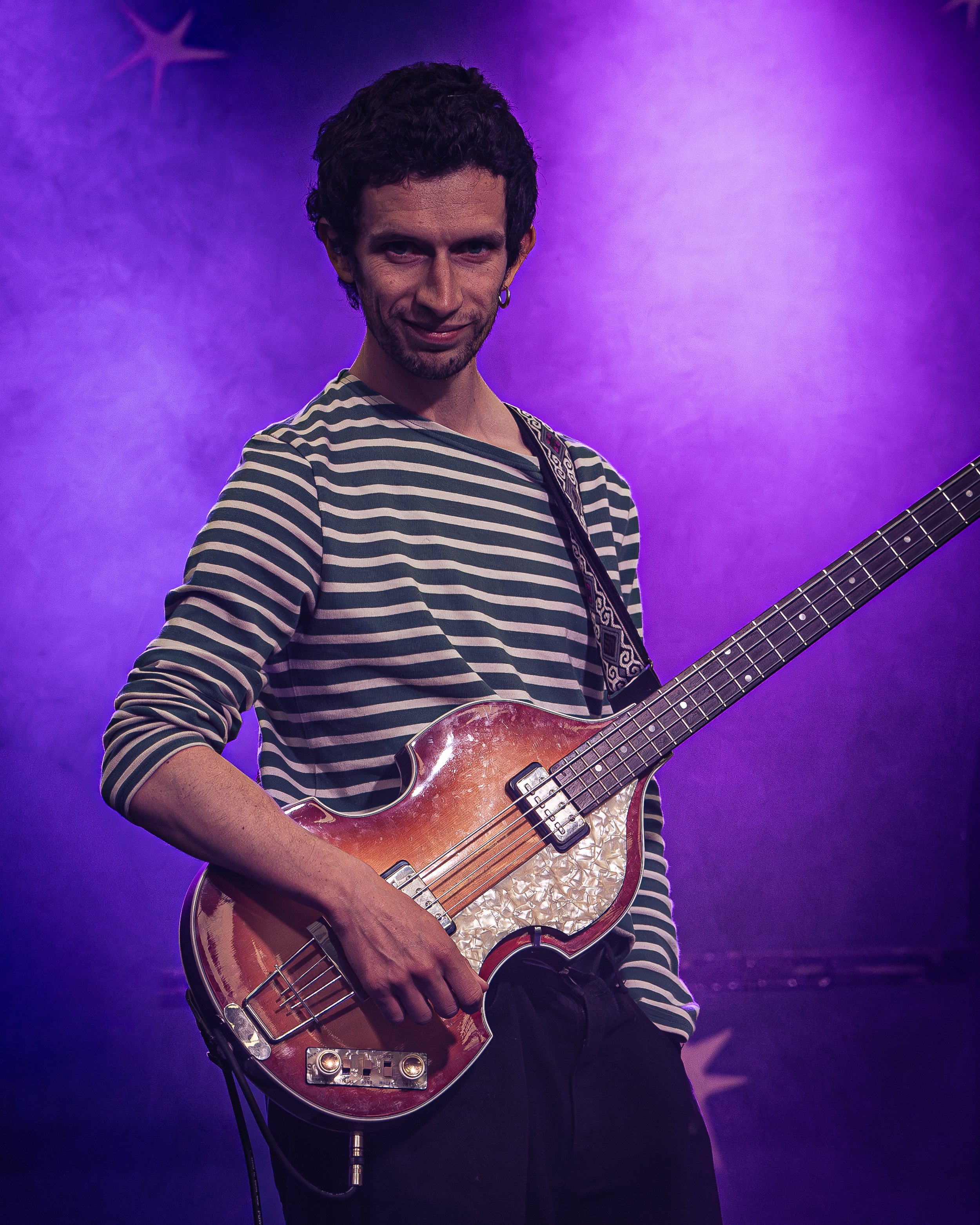 A man with dark curly hair, a beard, and a hoop earring in his left ear, wearing a black and white striped shirt, holding a guitar, standing against a purple-lit background with star-shaped decorations.