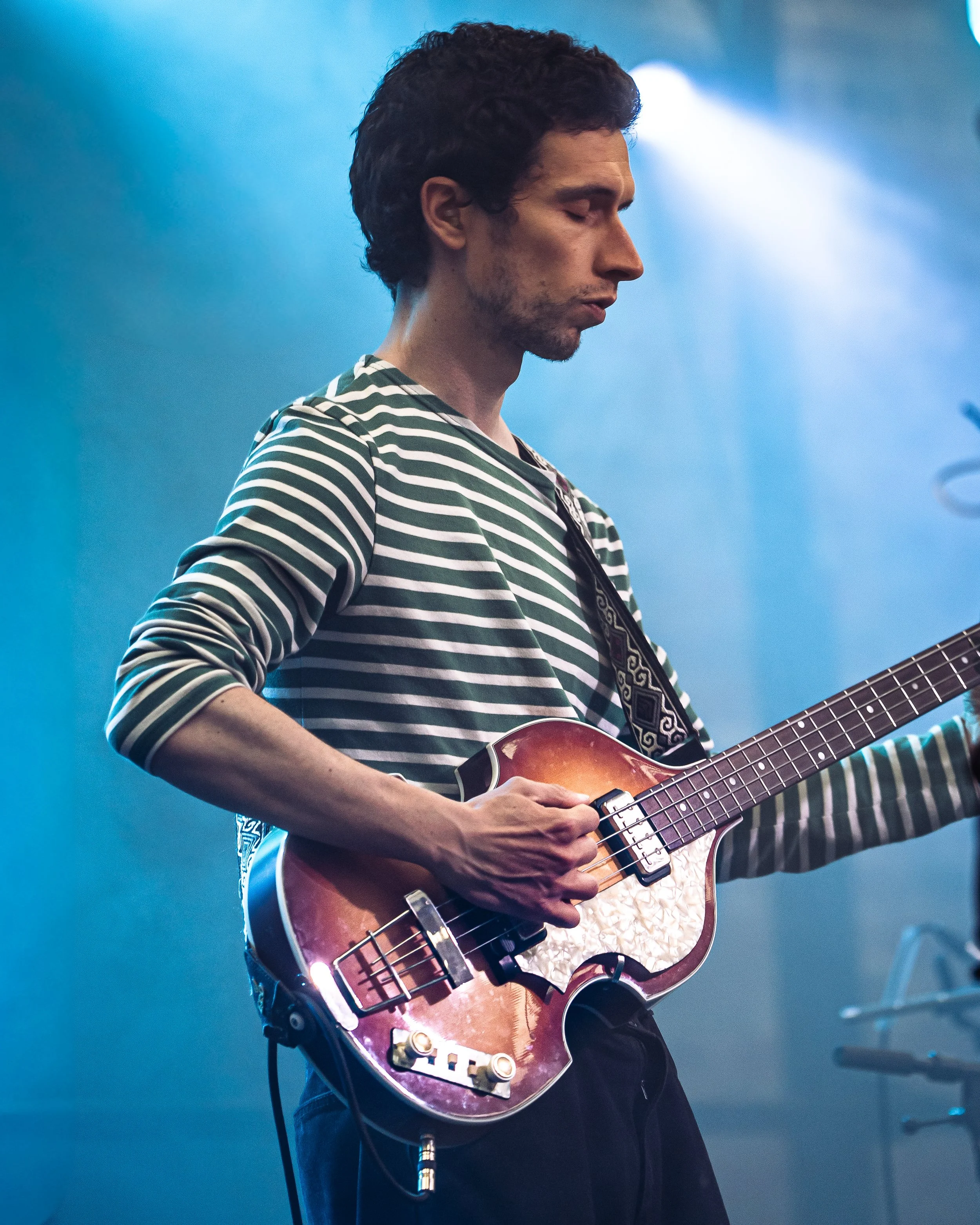 Man with dark hair and a striped shirt playing an electric guitar on stage with blue lighting.