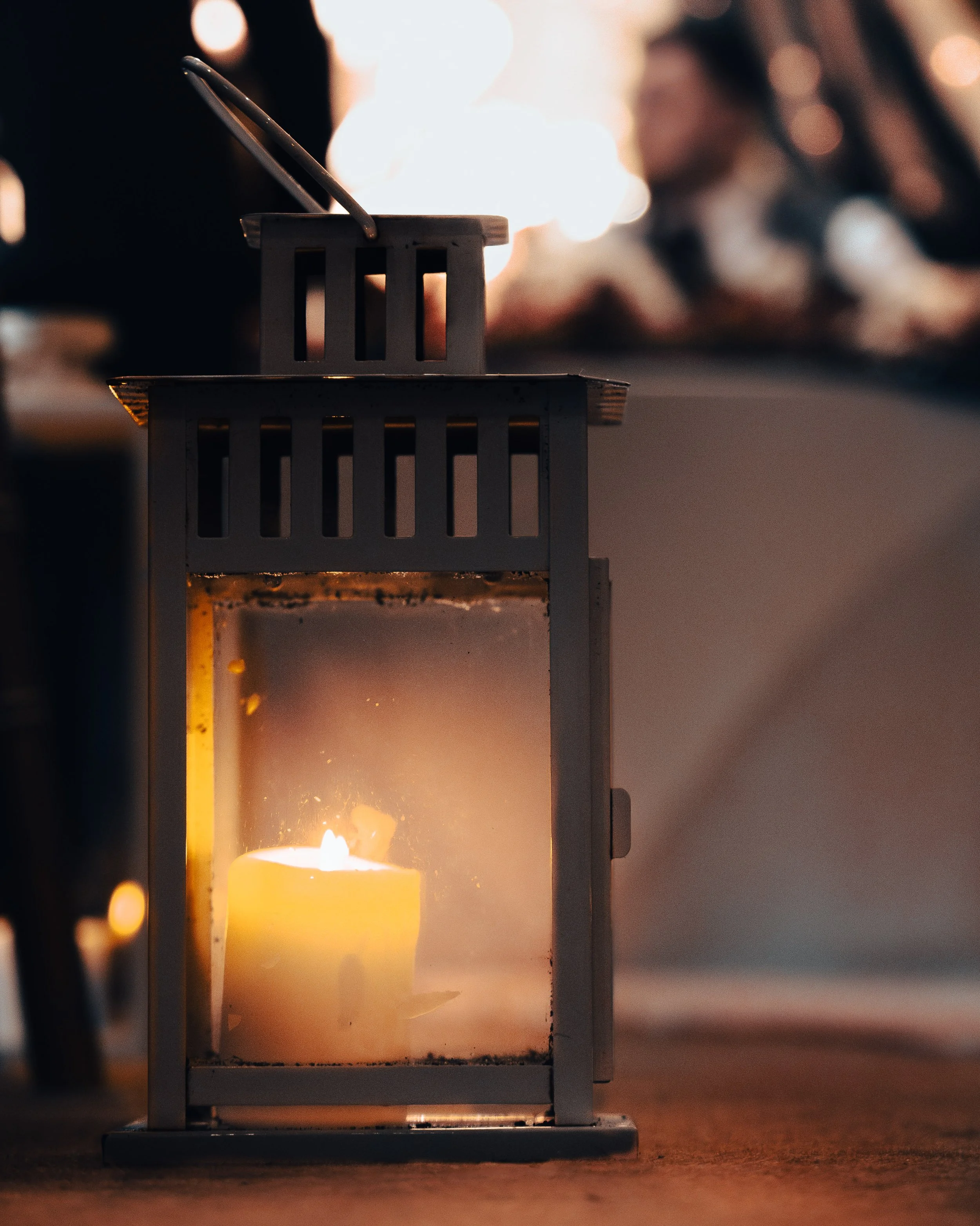 A lit candle inside a glass enclosure on a wooden surface, with a background of blurred warm lights and a person.