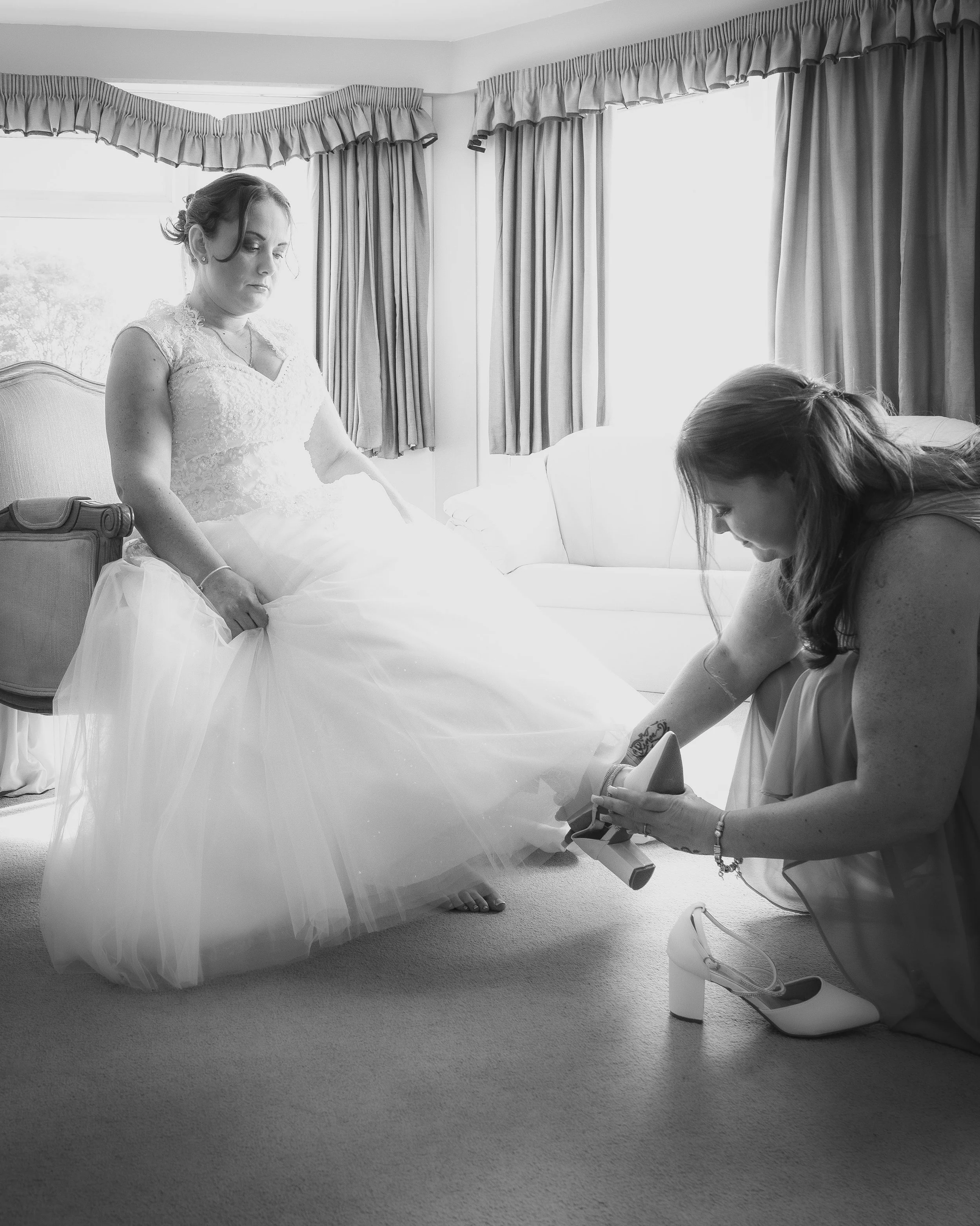 A bride sitting on a chair getting her wedding shoe adjusted by another woman in a room with natural light, curtains, and a sofa.