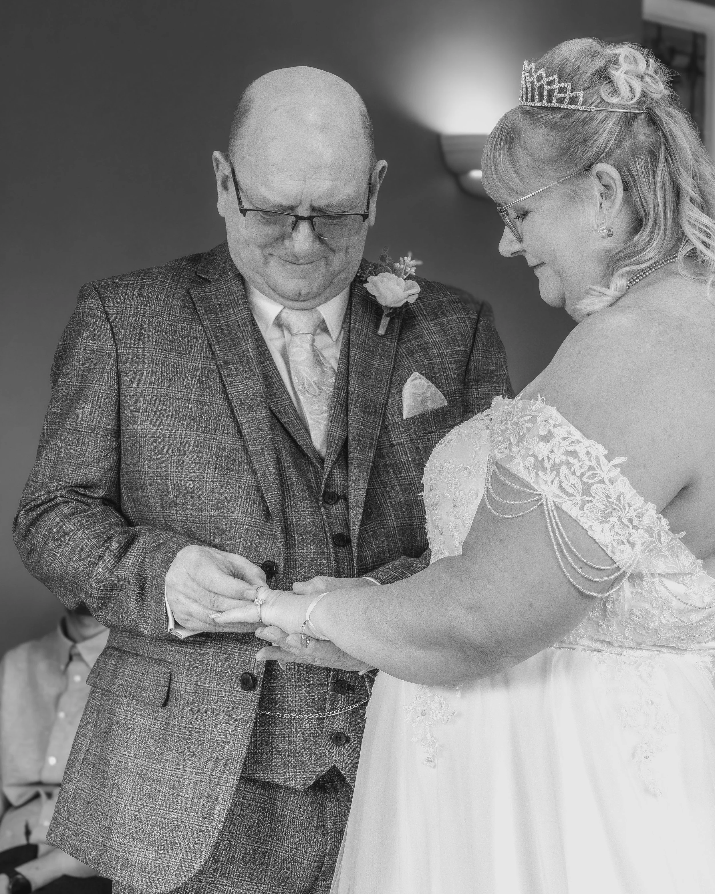 A black and white photograph of a wedding ceremony showing a groom placing a ring on the bride's finger. The groom is wearing glasses, a plaid suit with a boutonniere, and a tie. The bride is wearing a dress with lace and floral details, glasses, and