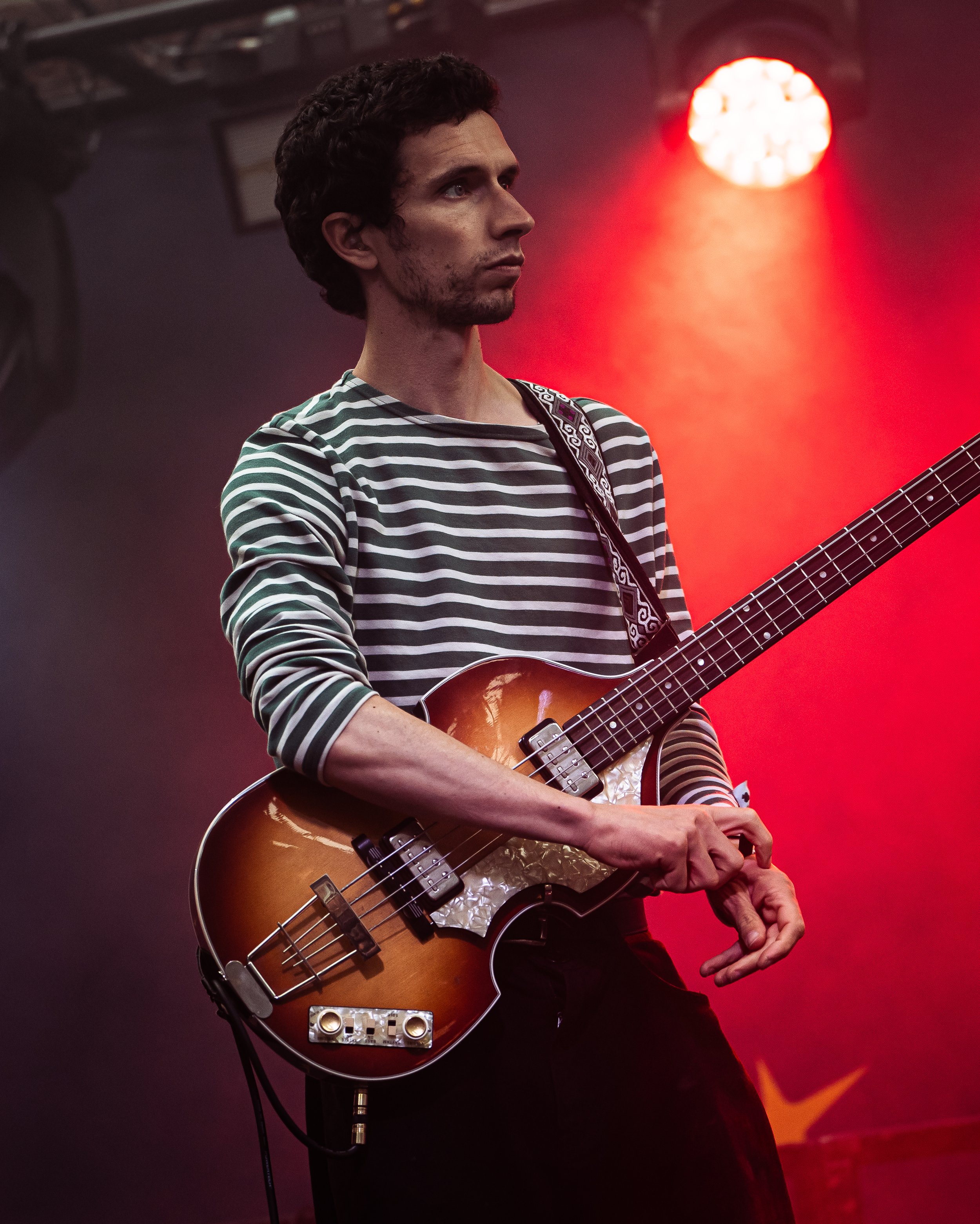 A man with curly hair and a casual striped shirt playing a sunburst electric bass guitar on a stage with red spotlight lighting.