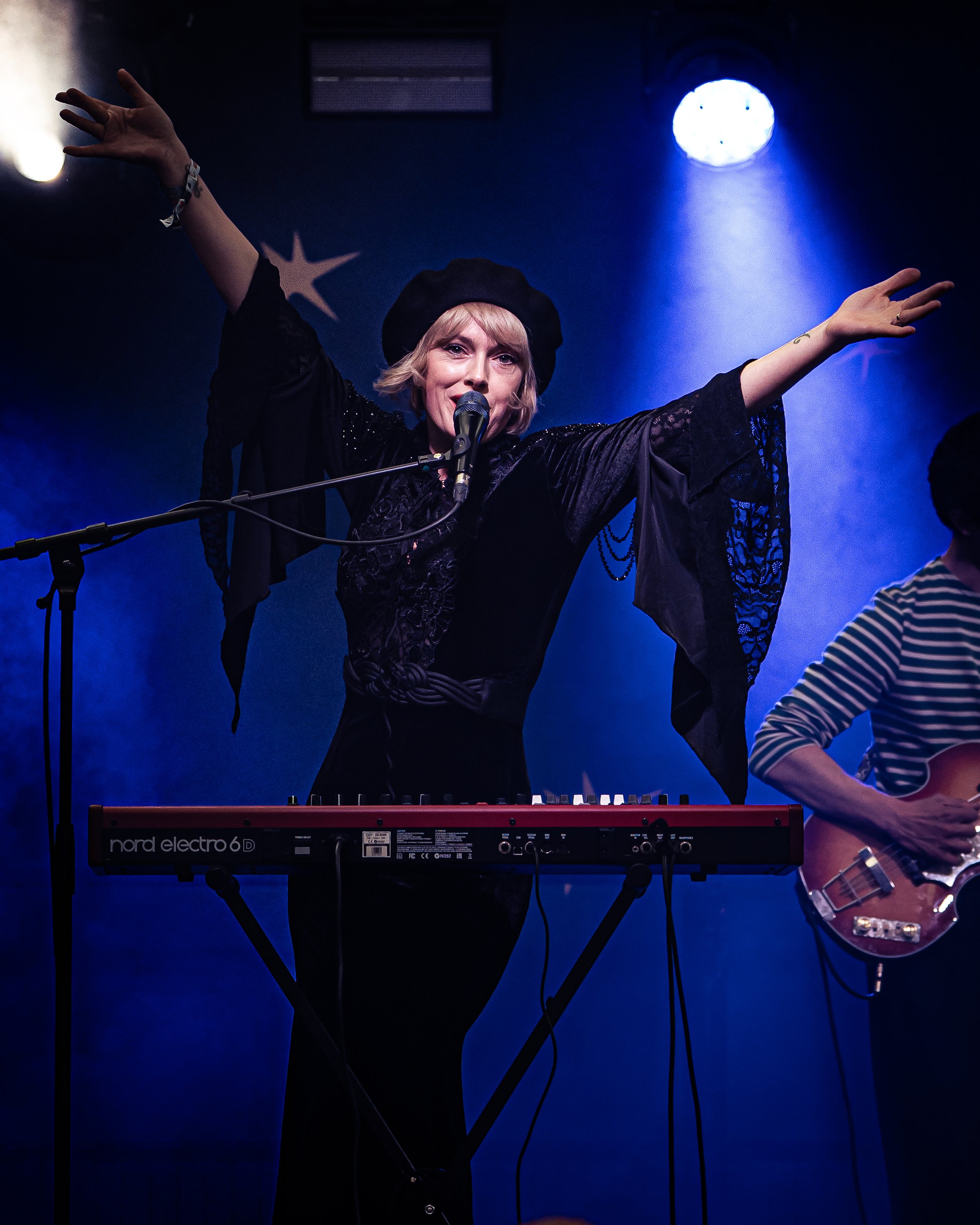 A woman with blonde hair, wearing a black beret and black lace dress, singing into a microphone on stage. She has her arms raised and is accompanied by a person playing a red electric guitar. The background features blue stage lighting and a large br