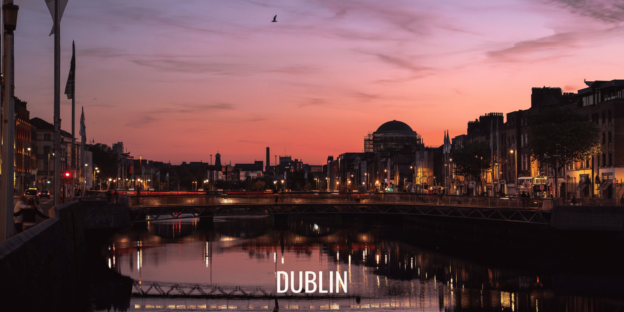 View of Dublin cityscape at dusk, with a river reflecting the illuminated buildings, trees, and a bridge, with a colorful pink, purple, and orange sky.