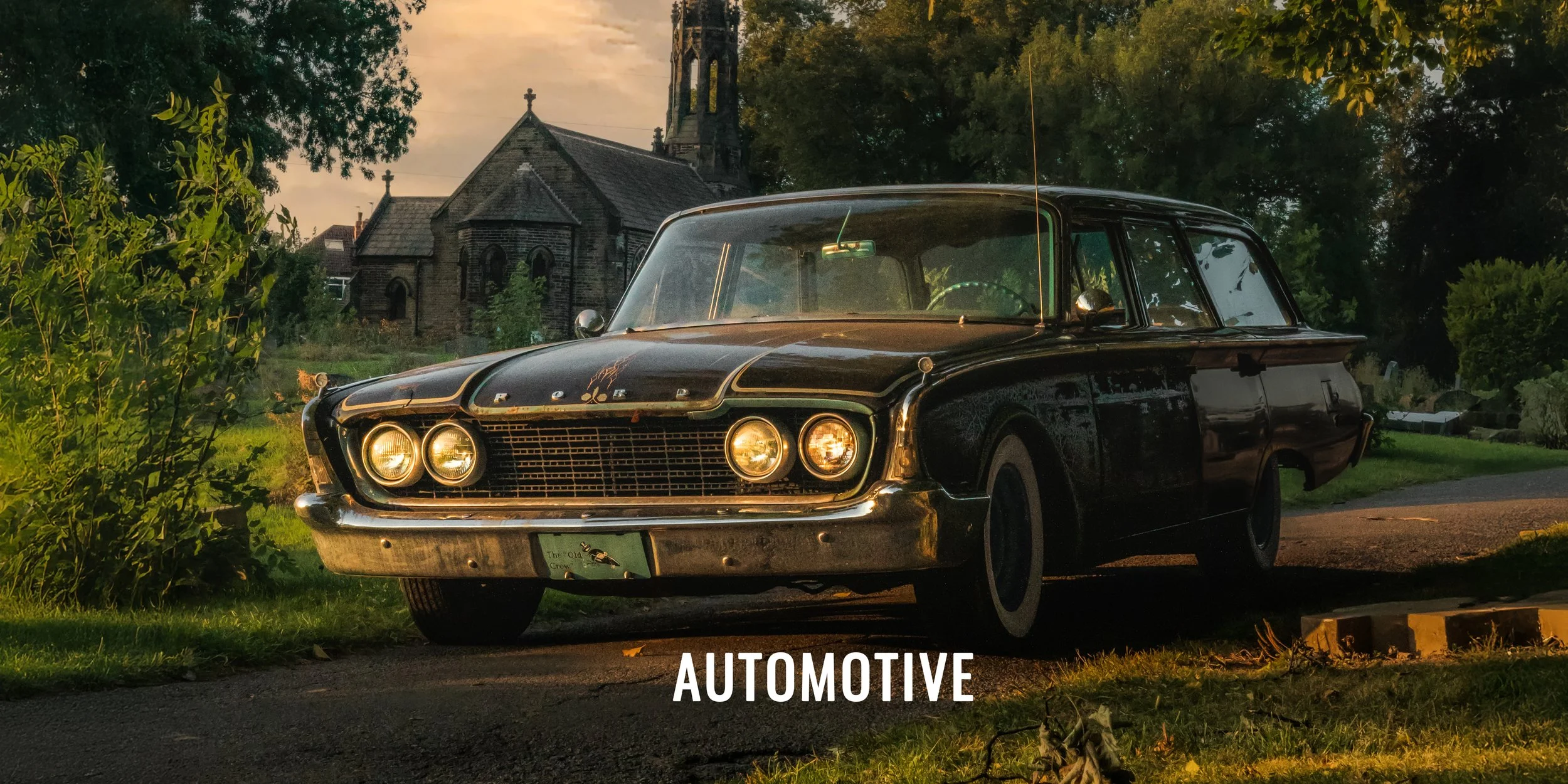 A vintage black station wagon car parked on the side of a road near greenery and a church in the background during sunset.