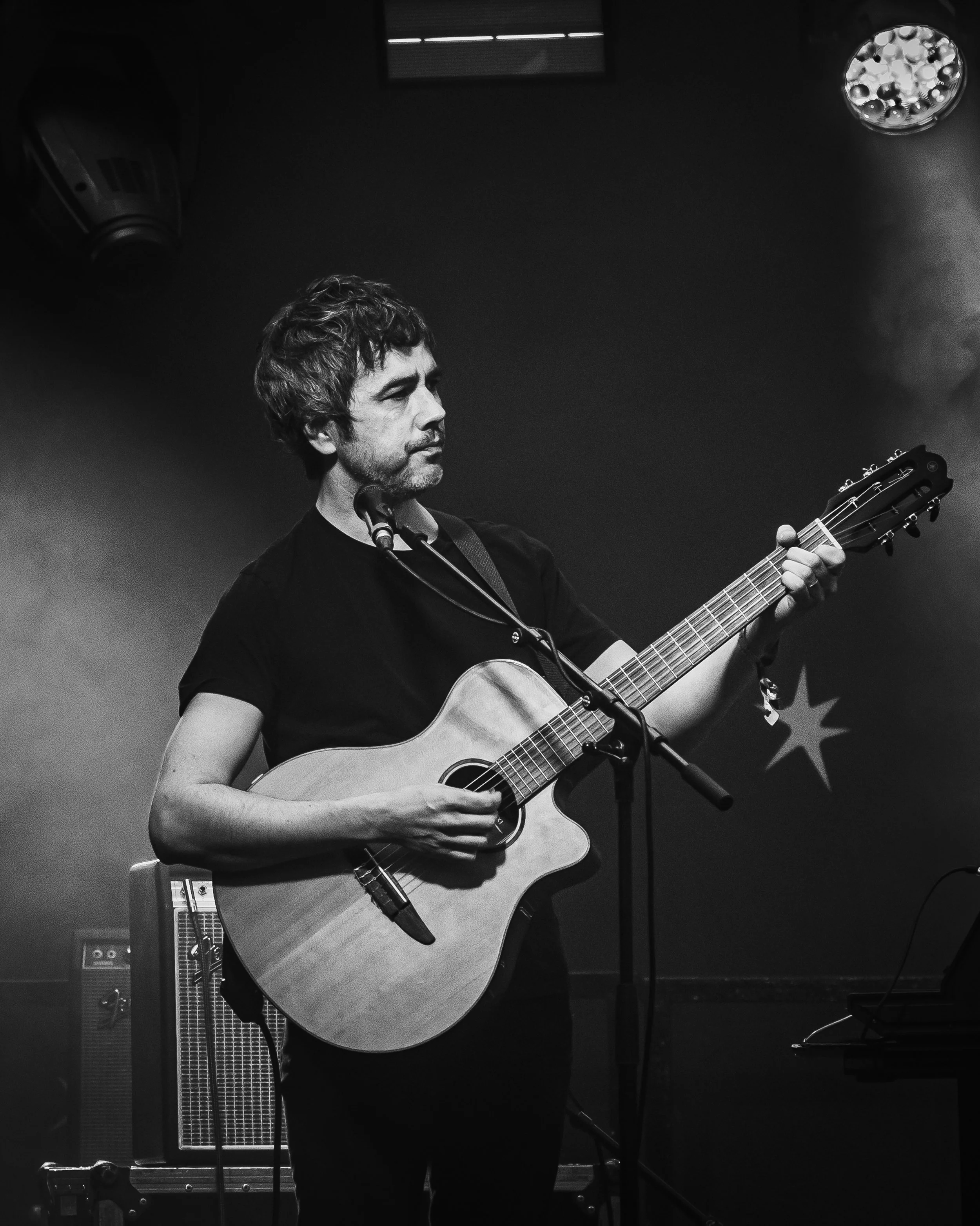 A man with dark hair and a beard playing an acoustic guitar on stage in a dimly lit setting, with a microphone in front of him, stage lights above, and musical equipment visible in the background.