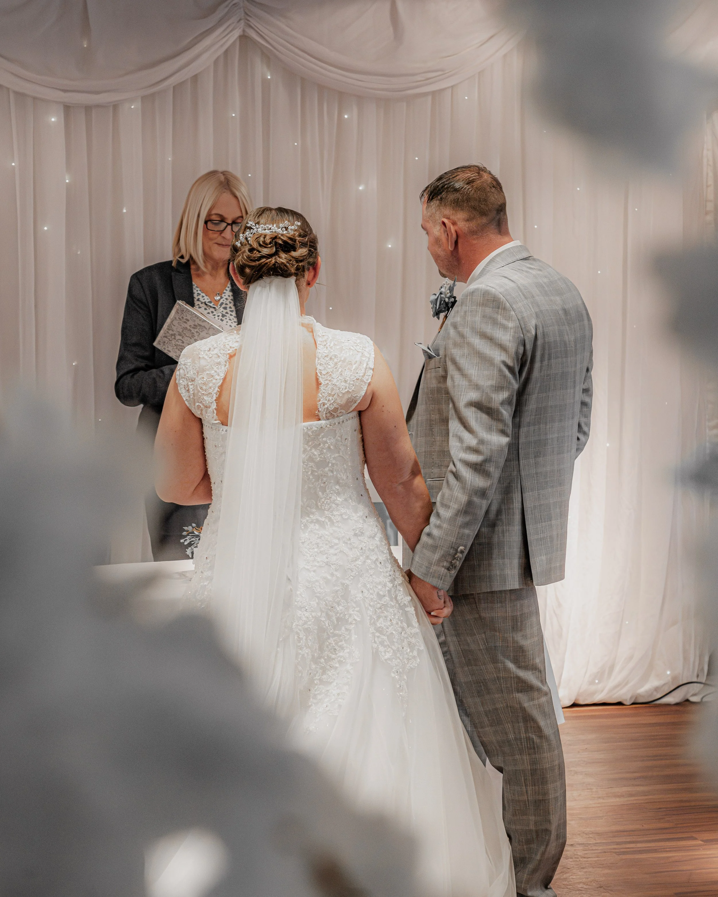 A couple holding hands during their wedding ceremony, facing an officiant, with a white curtain backdrop decorated with tiny lights.