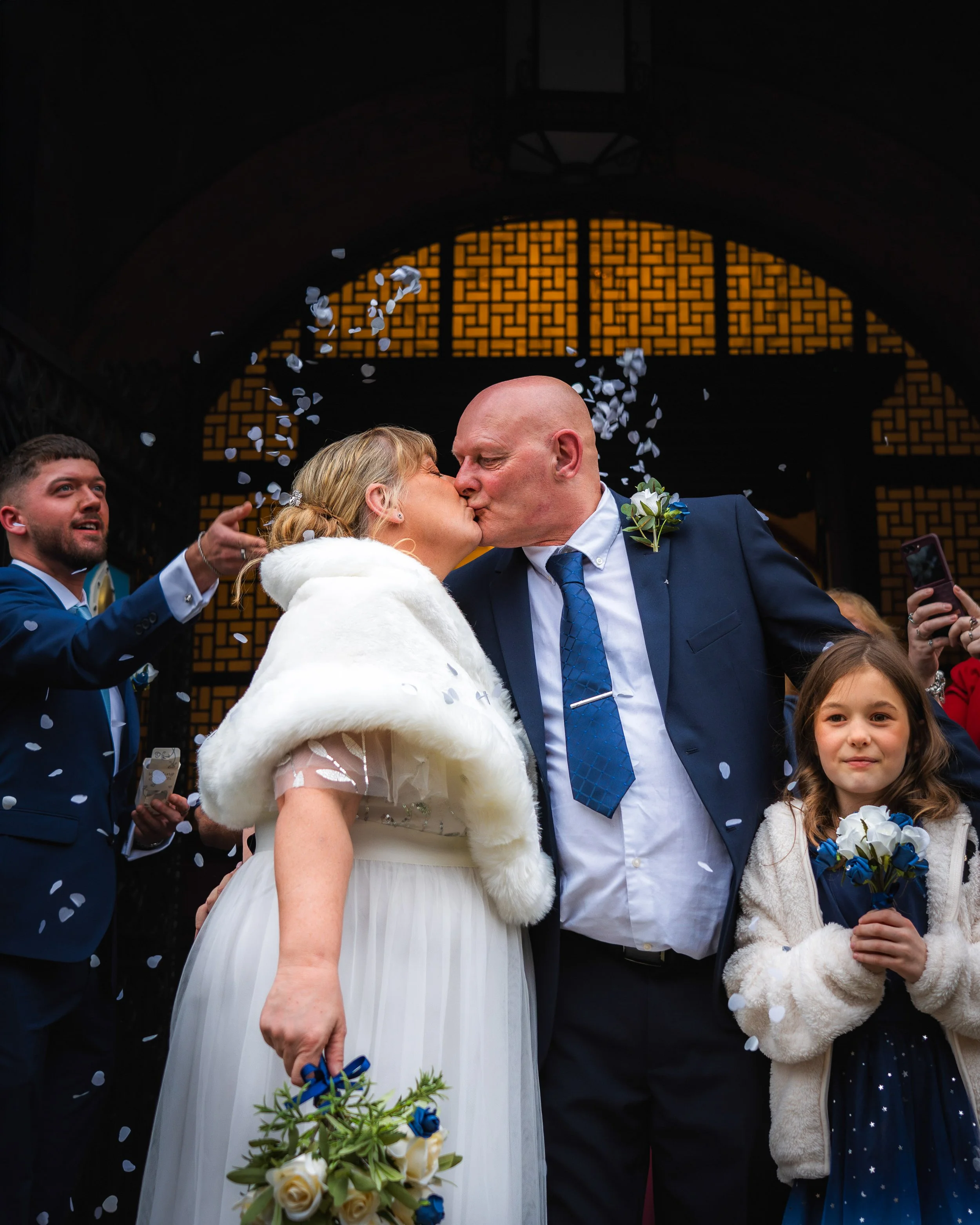 A bride and groom kiss during their wedding celebration, surrounded by guests throwing confetti inside a dark venue with a patterned orange background.
