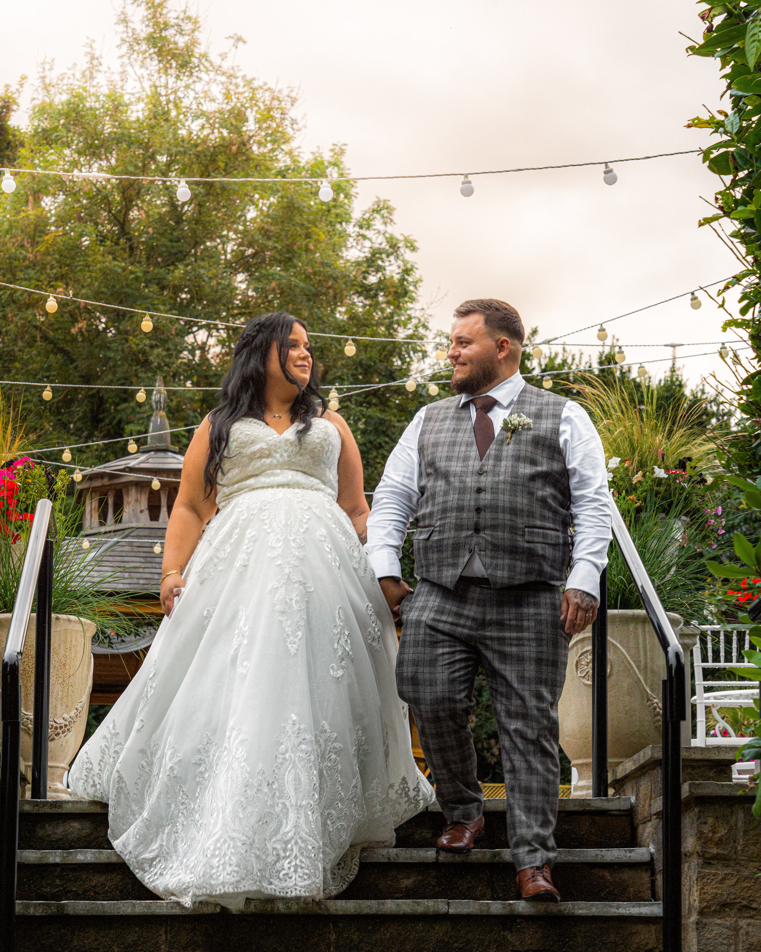 A bride and groom holding hands, walking down the steps outdoors during their wedding, surrounded by greenery and string lights.