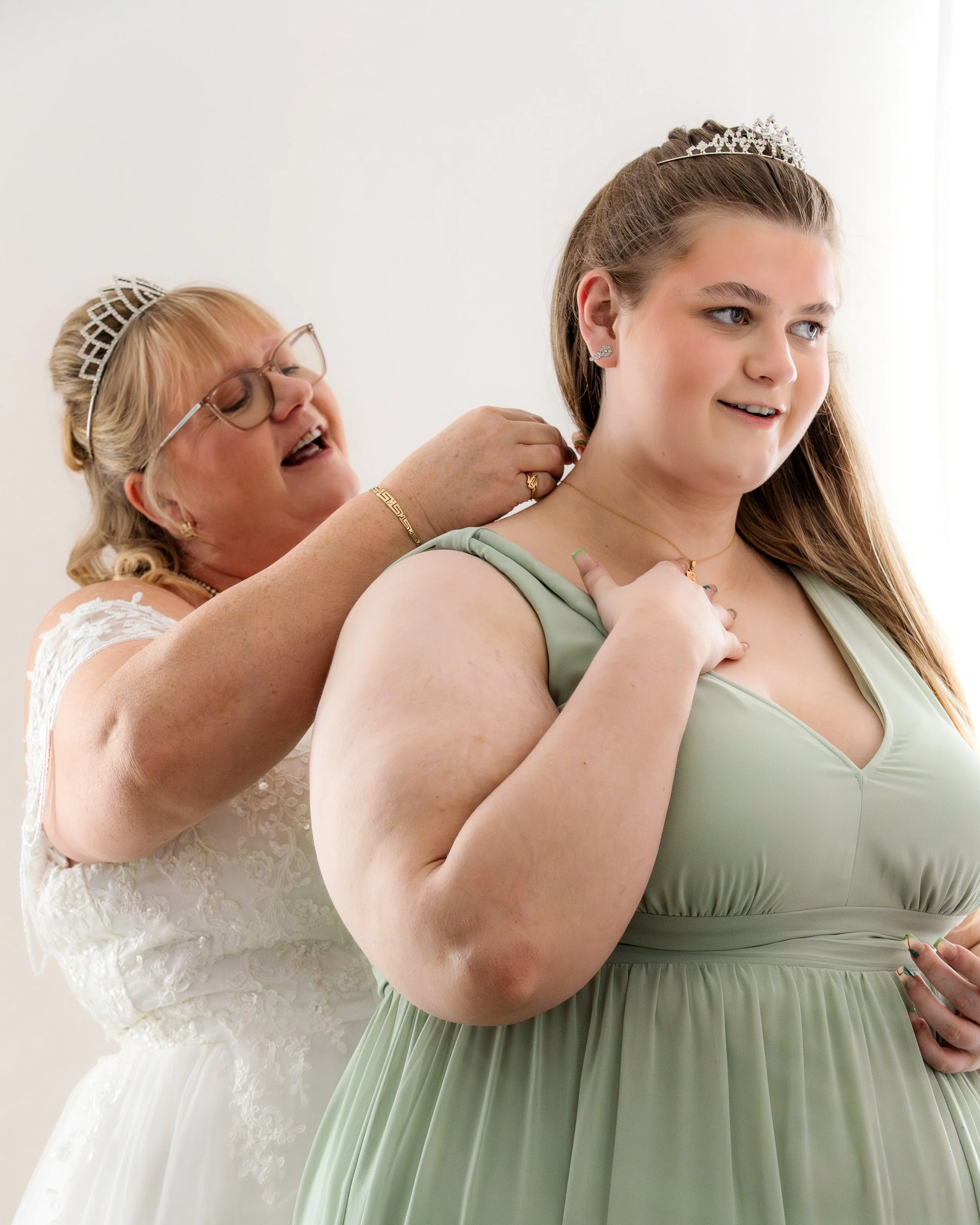 An older woman in a white dress and glasses helps a younger woman in a sage green dress put on a necklace. Both women are wearing tiaras.