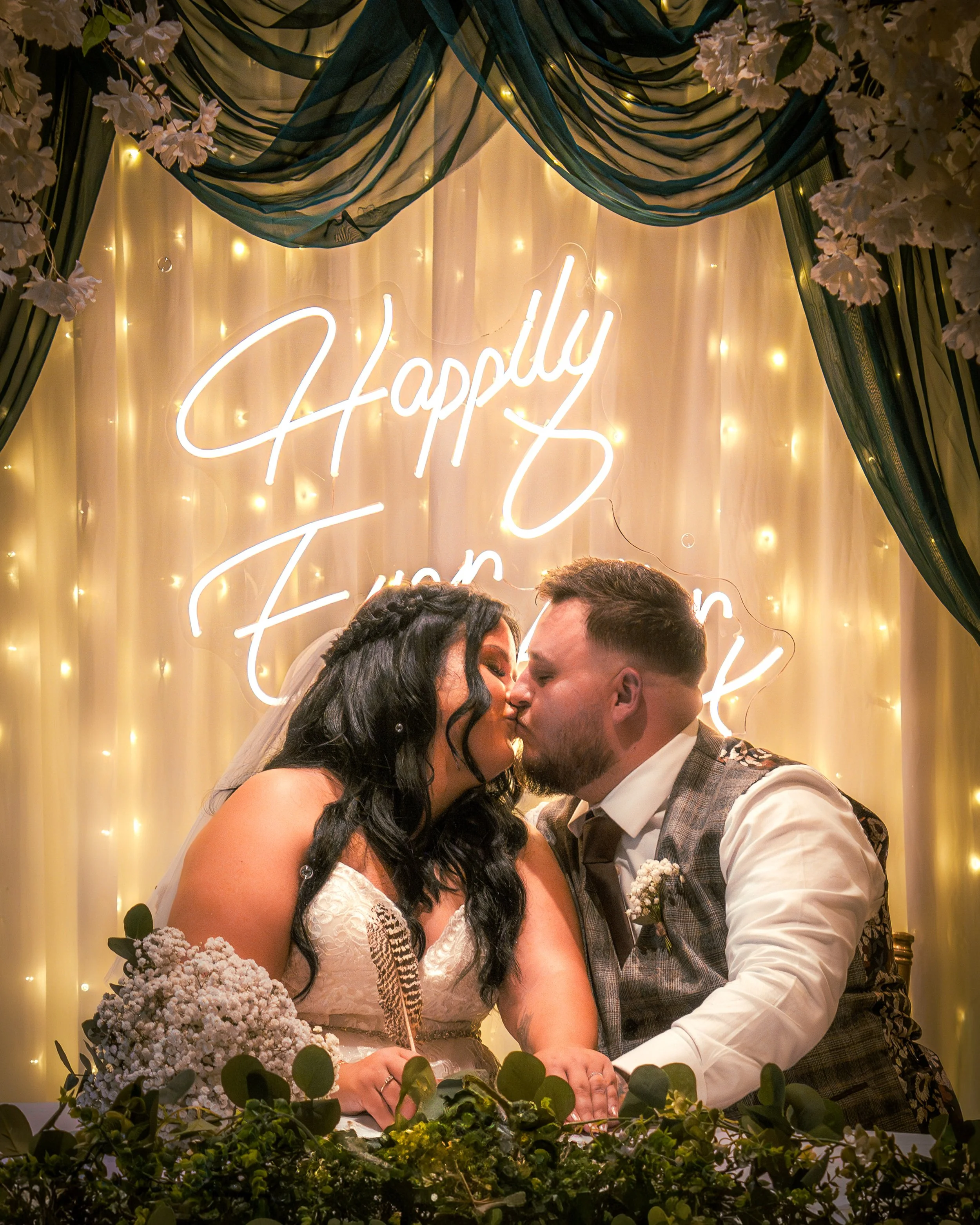 A newlywed couple sharing a kiss at their wedding reception, with a glowing sign that reads 'Happily Ever After' and decorated with fairy lights and greenery.