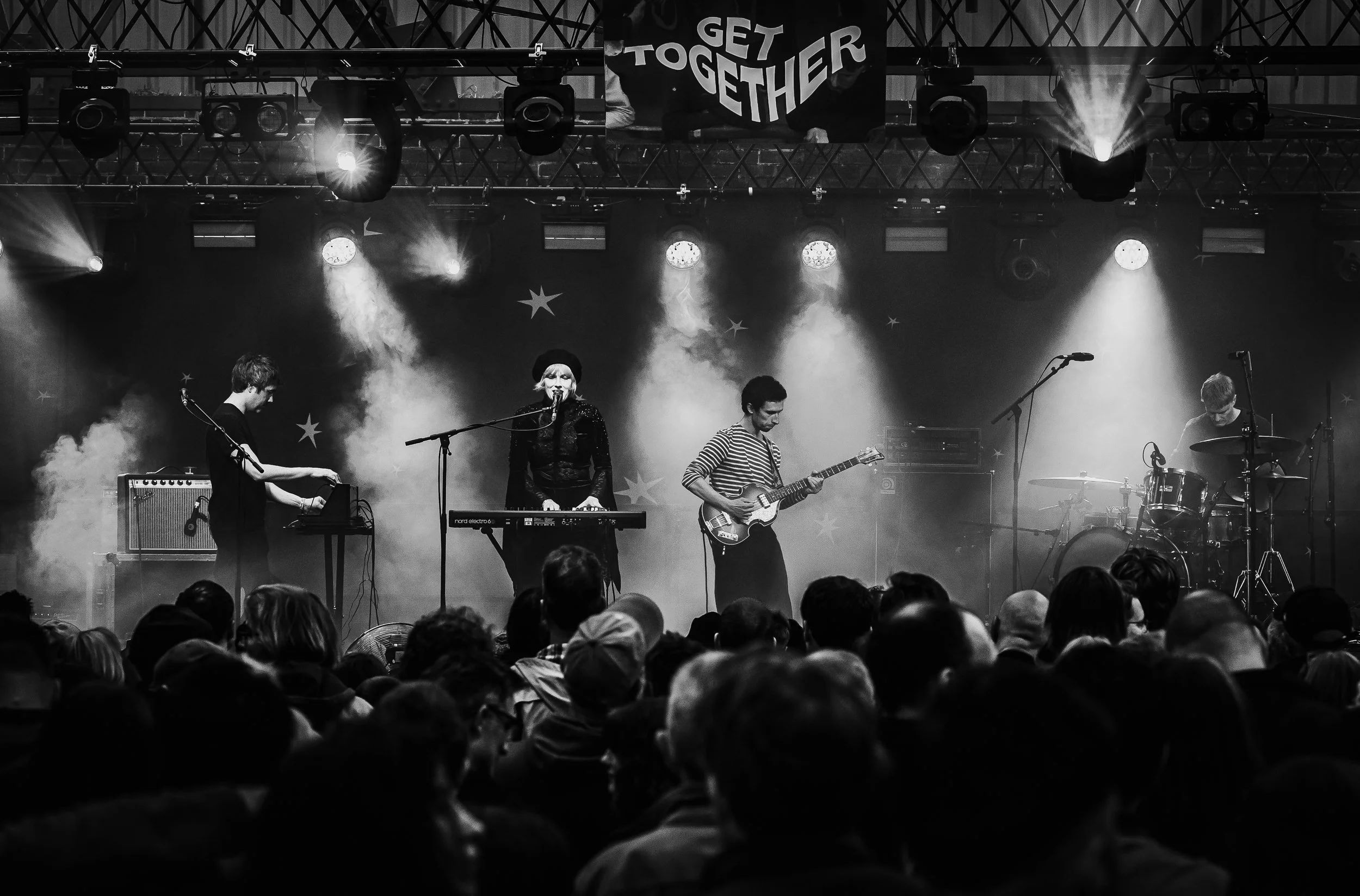 Black and white photo of a live band performing on stage with four musicians: a keyboard player, a singer, a guitarist, and a drummer. The stage has lighting effects and a banner above with the words 'GET TOGETHER'.