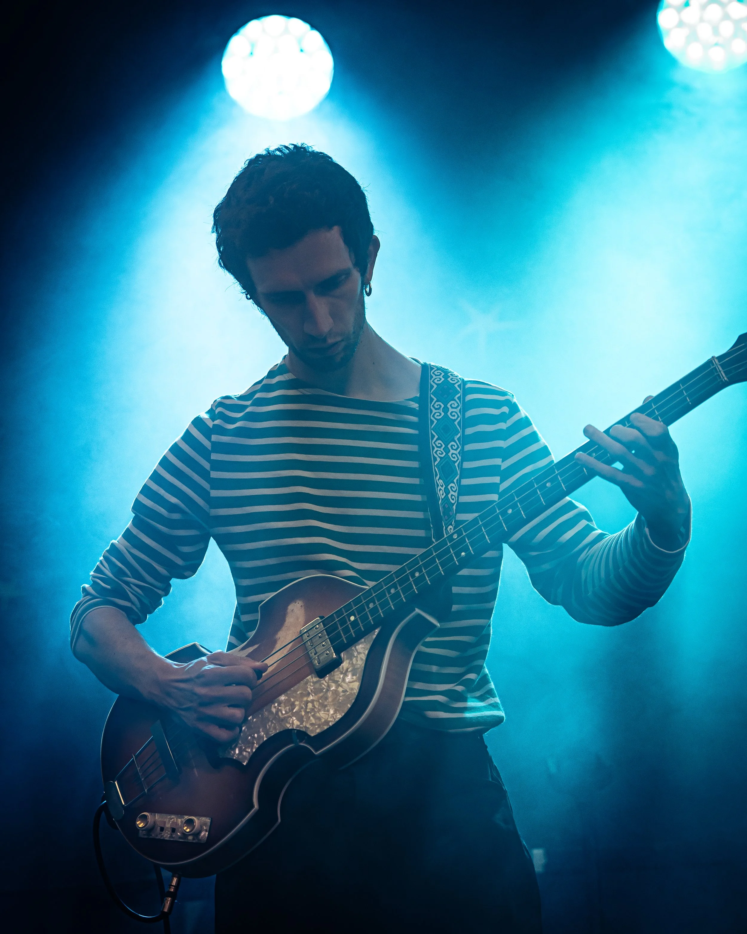 A musician in a striped shirt playing an electric guitar on stage with blue lighting.