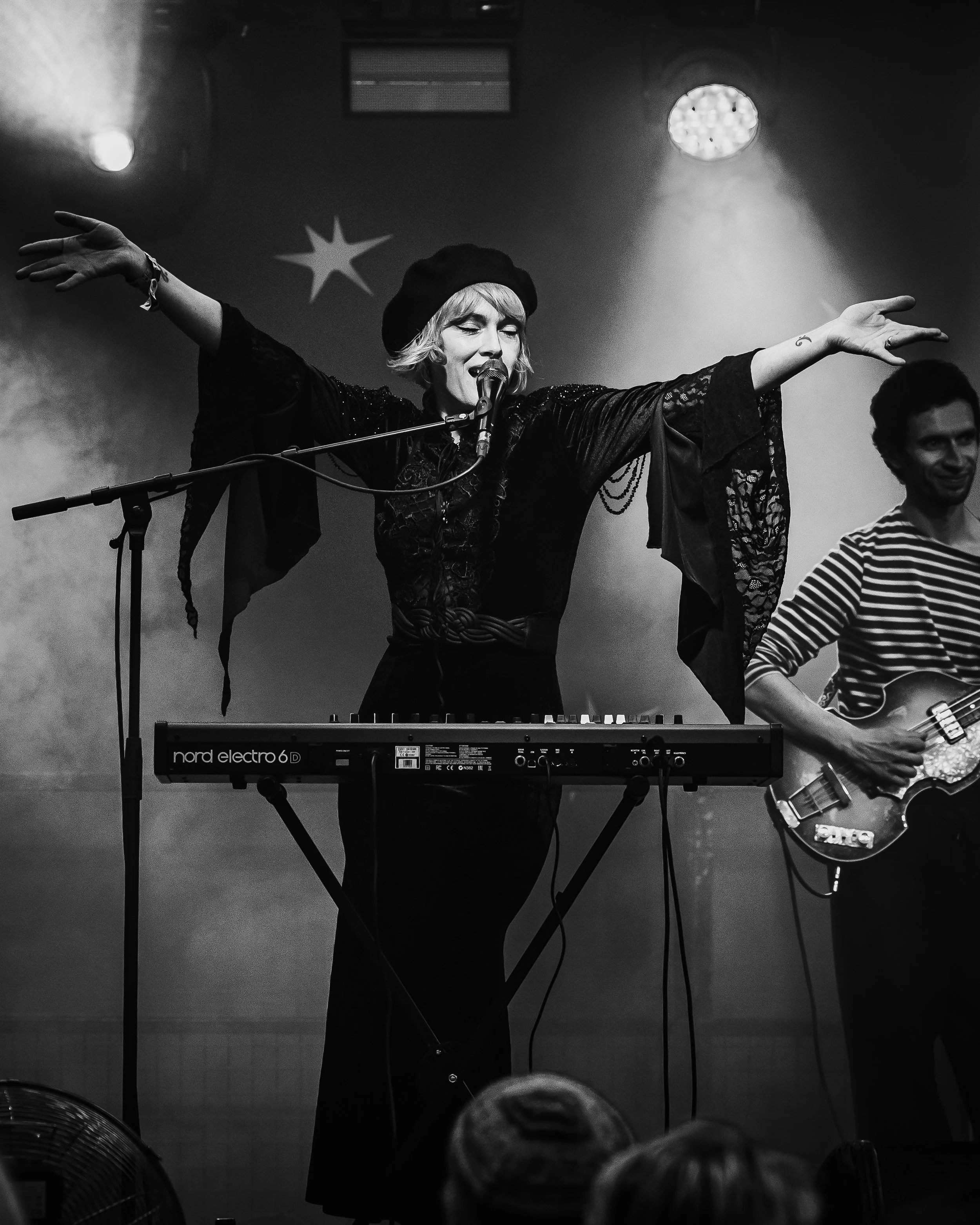 Black and white photo of a woman singing into a microphone with her arms outstretched, playing a keyboard in front of her, and a man playing guitar in the background, in a live music venue.
