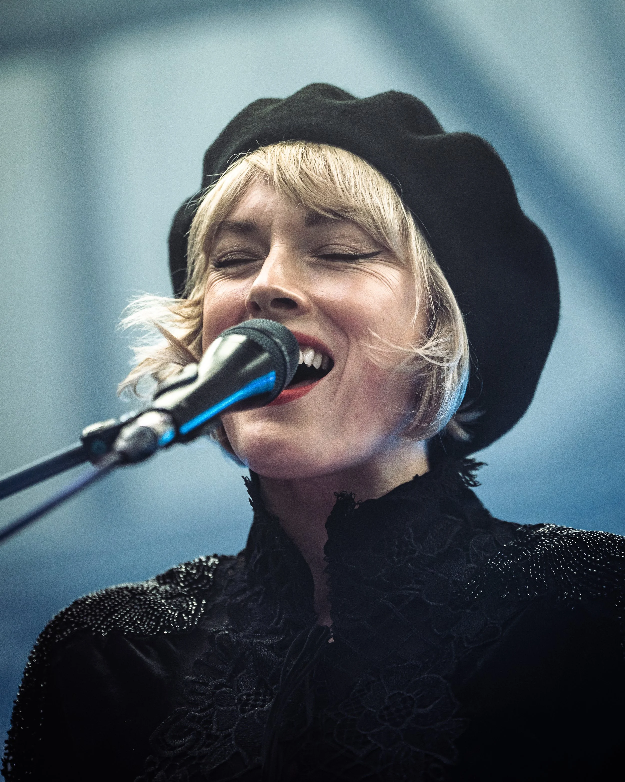 A woman with light blonde hair, smiling with eyes closed, wearing a black hat and black textured lace top, speaking or singing into a microphone.
