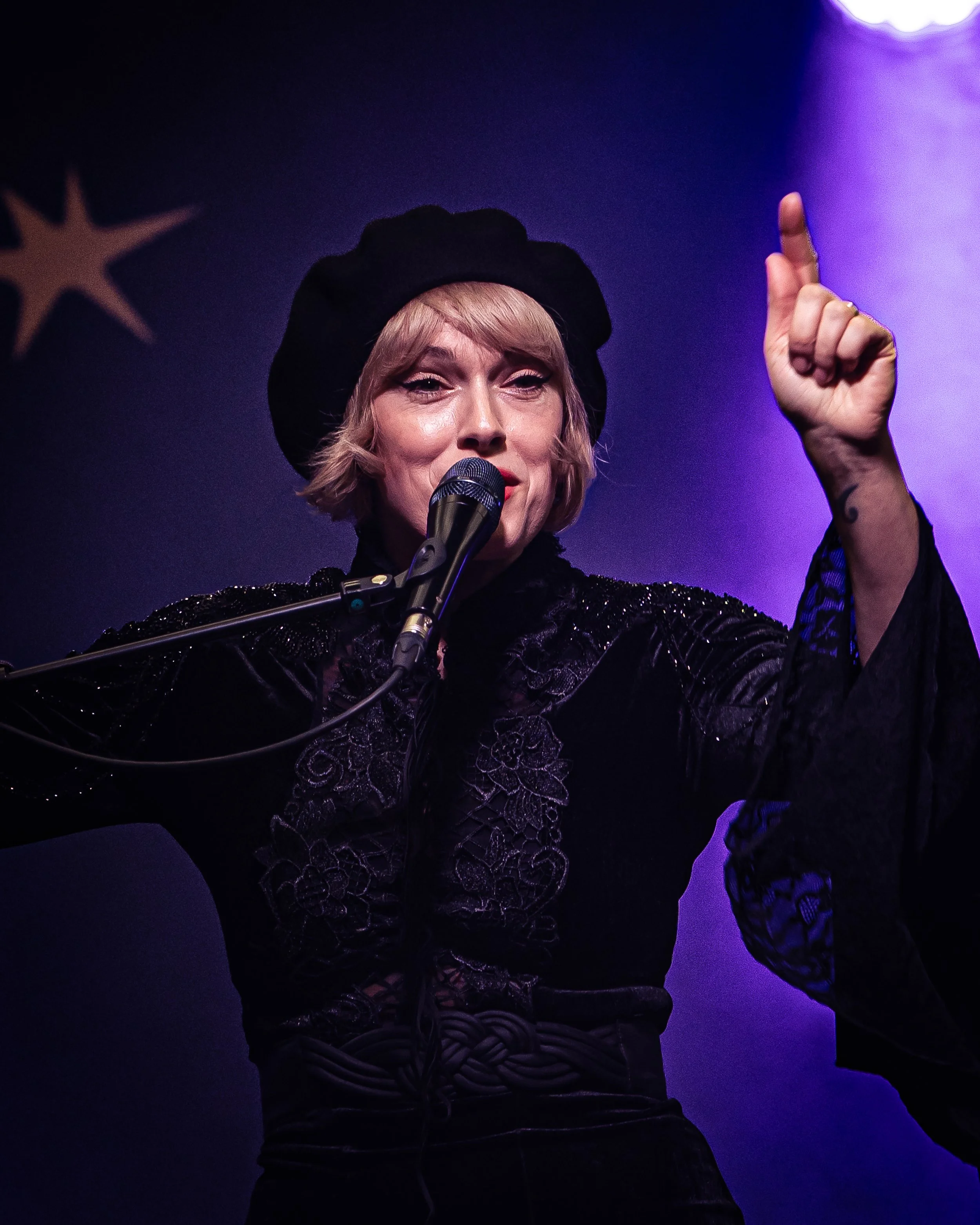 A woman singing into a microphone on stage, wearing a black hat, black outfit with lace details, and raising her hand with a finger pointing upward, purple stage lighting behind her.