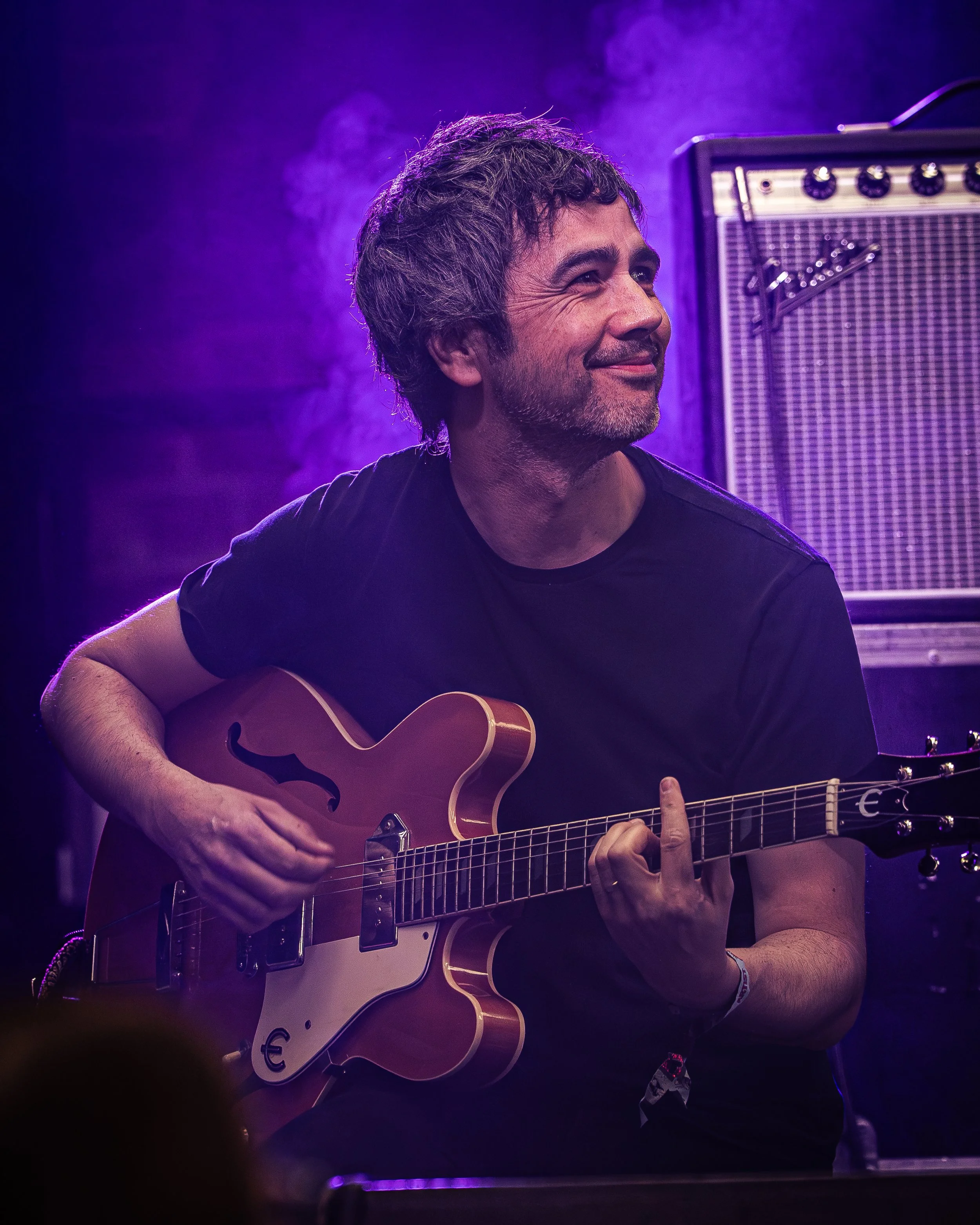 A man with dark, curly hair and a beard playing an electric guitar on stage under purple lighting.