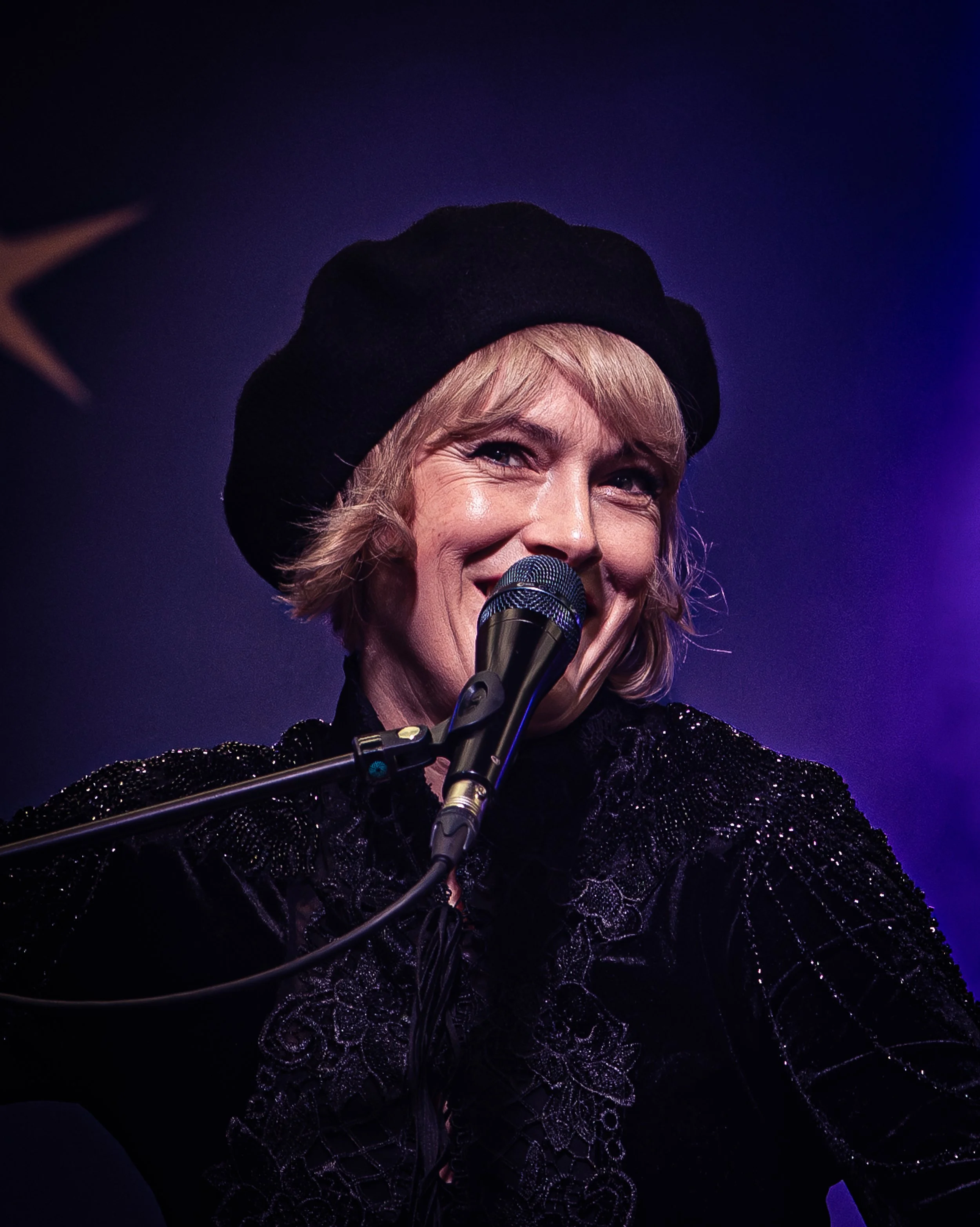 A woman smiling and speaking into a microphone on stage, wearing a black beret and a black, metallic, lace dress with intricate floral patterns, with stage lighting in the background.