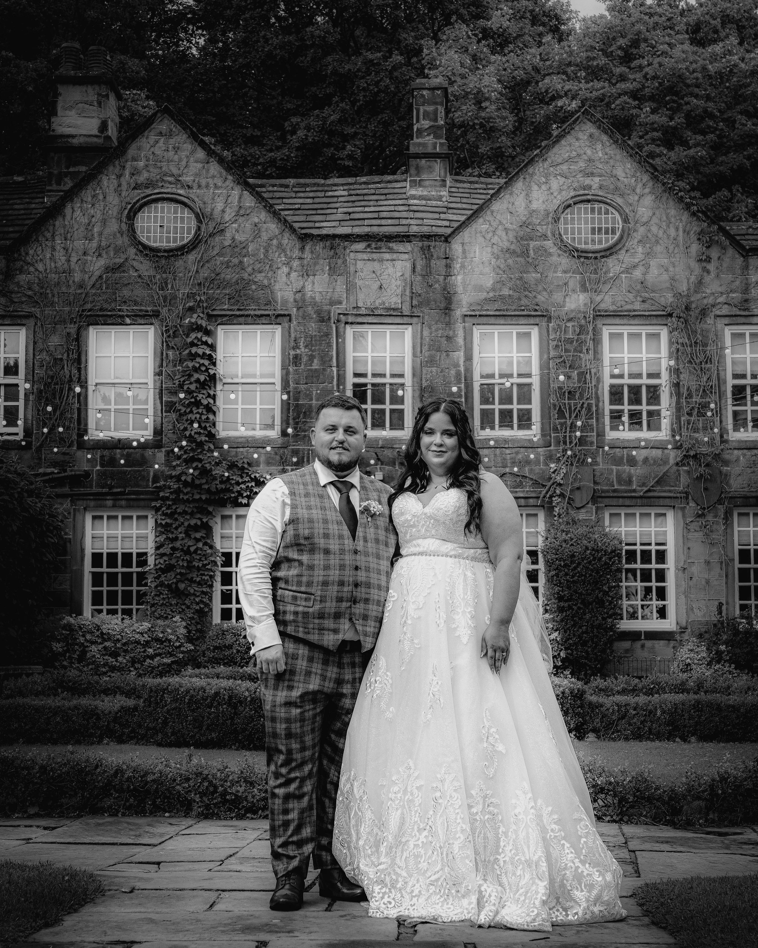 Black and white photo of a bride and groom standing in front of an old stone house with multiple windows and ivy growing on its walls, outdoors with trees in the background.