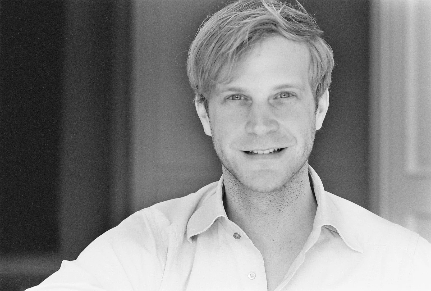 Black and white portrait of a smiling man with light-colored hair, wearing a collared shirt.